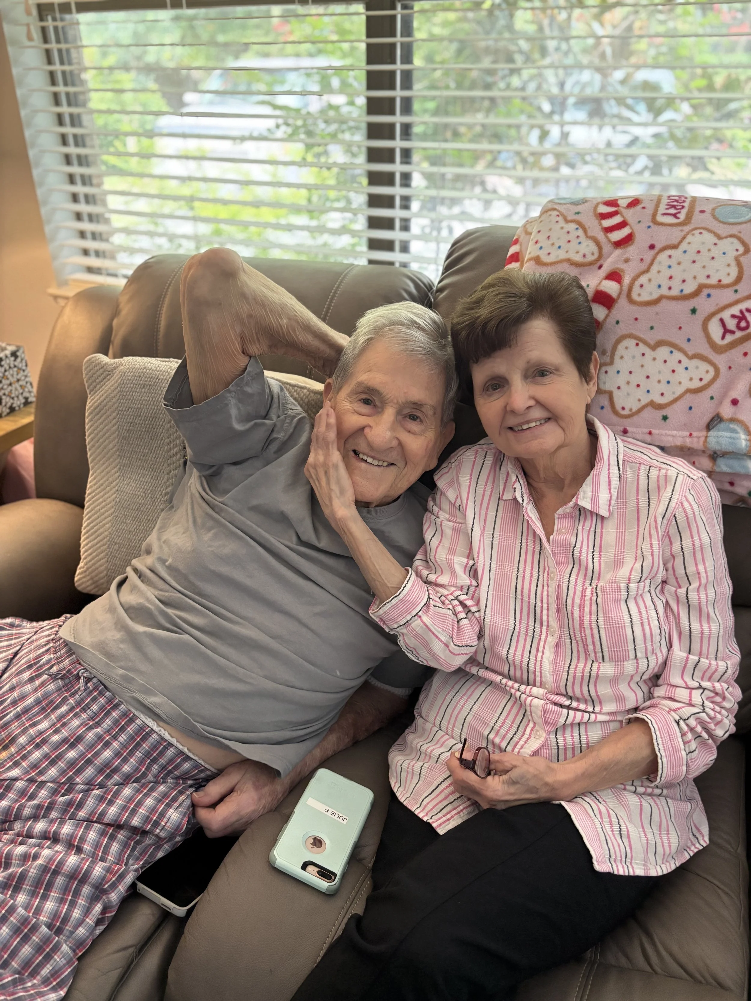 An elderly man and woman are sitting on a beige couch in a living room. The man is leaning back with his hand behind his head, smiling. The woman is sitting beside him, smiling, holding a pair of glasses. There is a pink hair towel with holiday patte