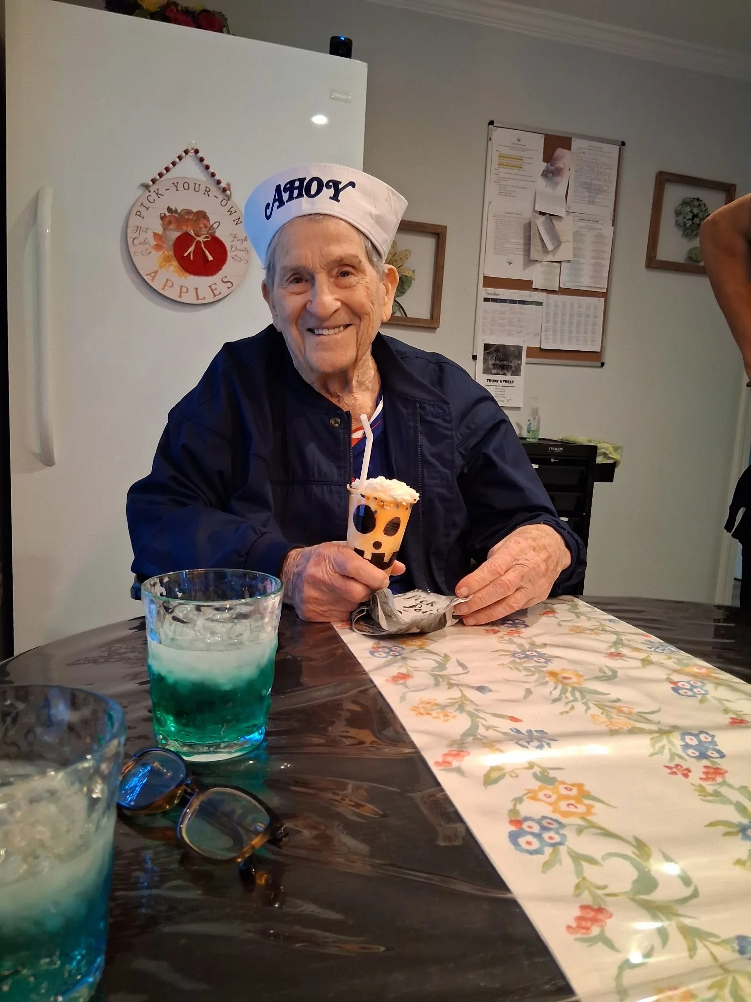 An elderly man wearing a paper chef's hat that reads "YAH" and a dark jacket, smiling and holding a pumpkin-shaped drink with a straw, seated at a table with glasses of colorful drinks, on a floral tablecloth inside a room decorated with a wall plaqu