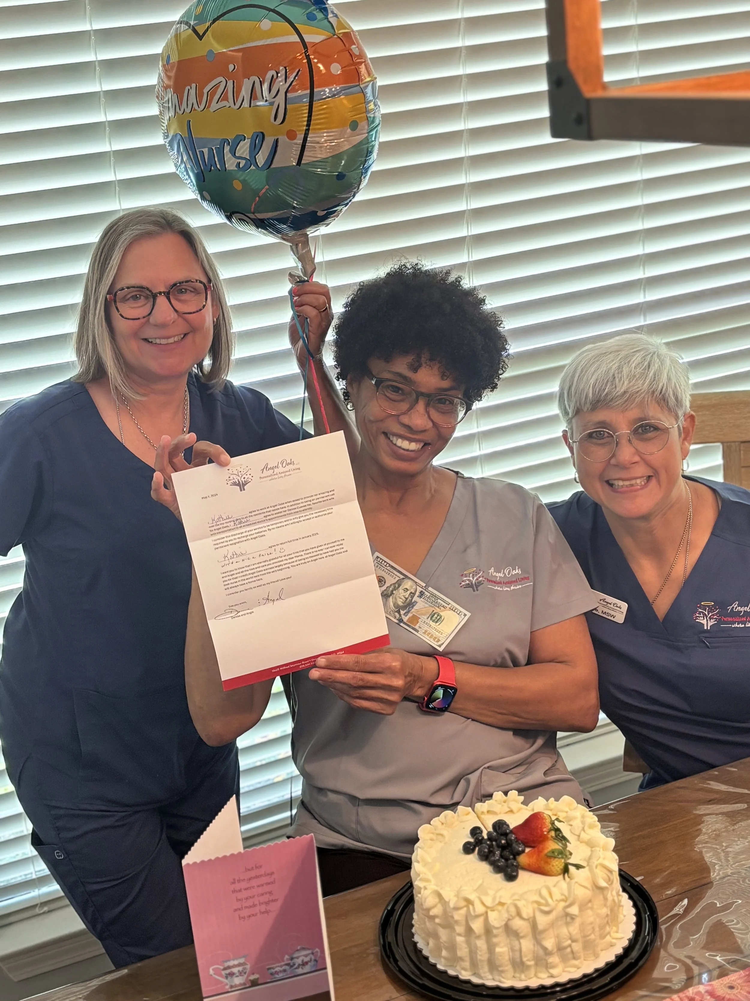 Three women celebrating in front of a cake with fruit toppings at a gathering, with one woman holding a colorful balloon that says 'amazing nurse' and a certificate.