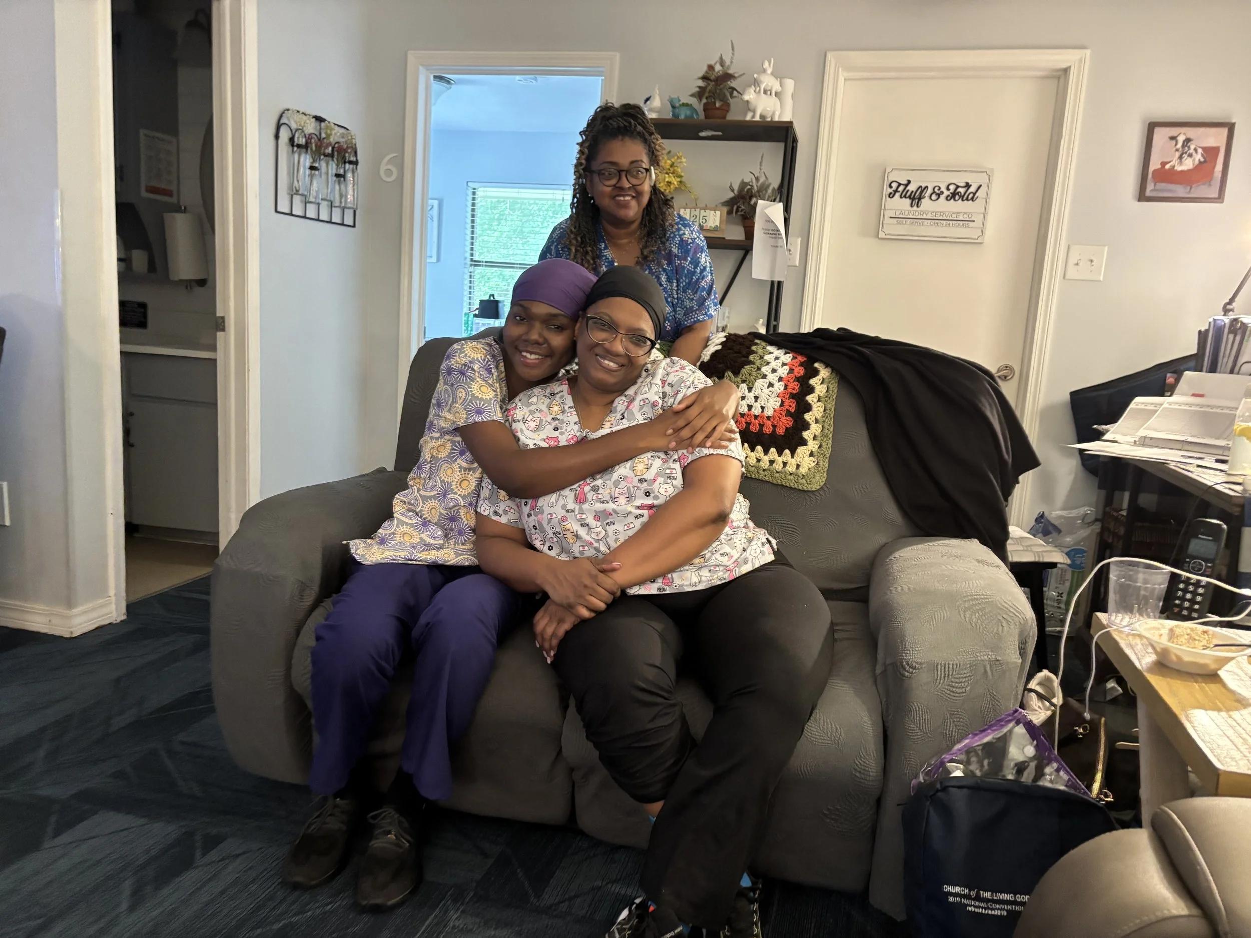 Three women are sitting and standing on a couch in a living room, smiling and posing for the photo. They are wearing casual clothing, and the woman in the middle has her hand on the arm of the woman in front. The room has a doorway with a window behi
