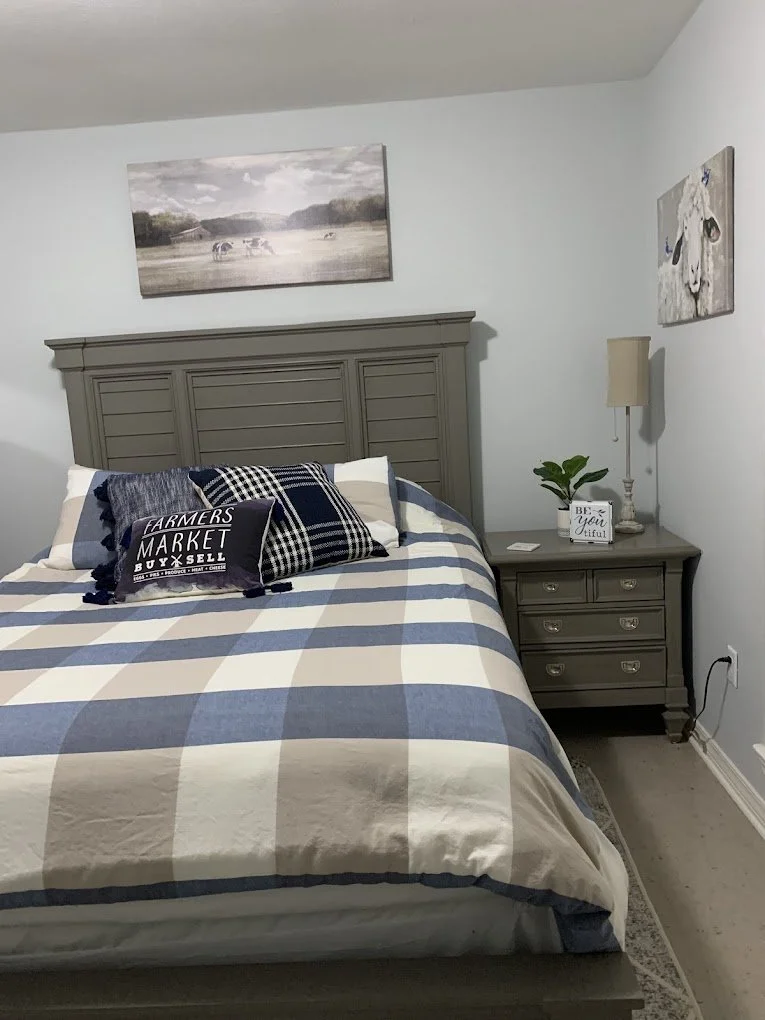 A bedroom with a grey wooden headboard, a quilted bed with checkered pillows, a nightstand with a plant, a lamp, and a decorative sign reading 'Be your beautiful.' Artwork of dogs hangs on the wall.