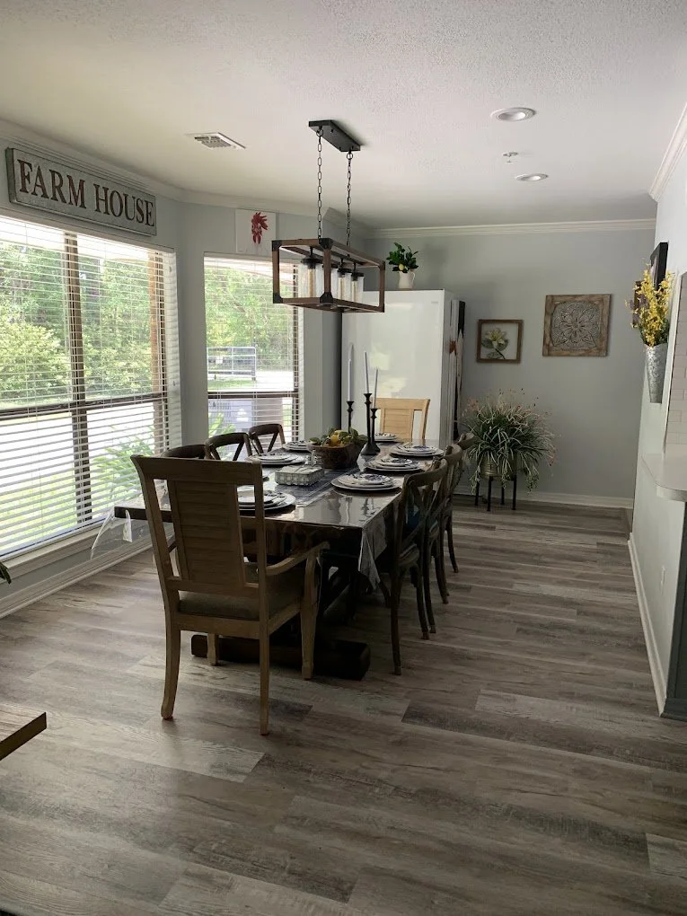 A dining room with a dark wooden table, eight chairs, a centerpiece with fruit, and a black candelabra. Large windows with blinds, light-colored walls, and a wooden floor. Decor includes a "Farm House" sign, wall art, and plants.