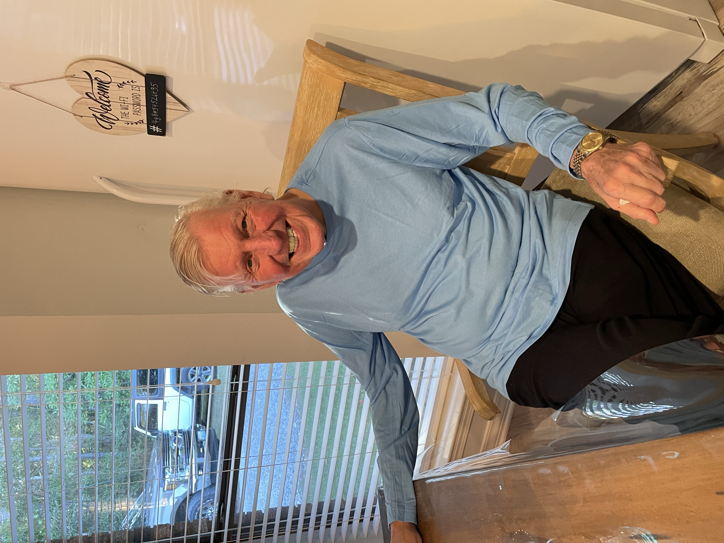An older man with white hair smiling while sitting in a wooden chair in a room with a window showing a parking lot outside. He is wearing a light blue shirt and a gold watch.