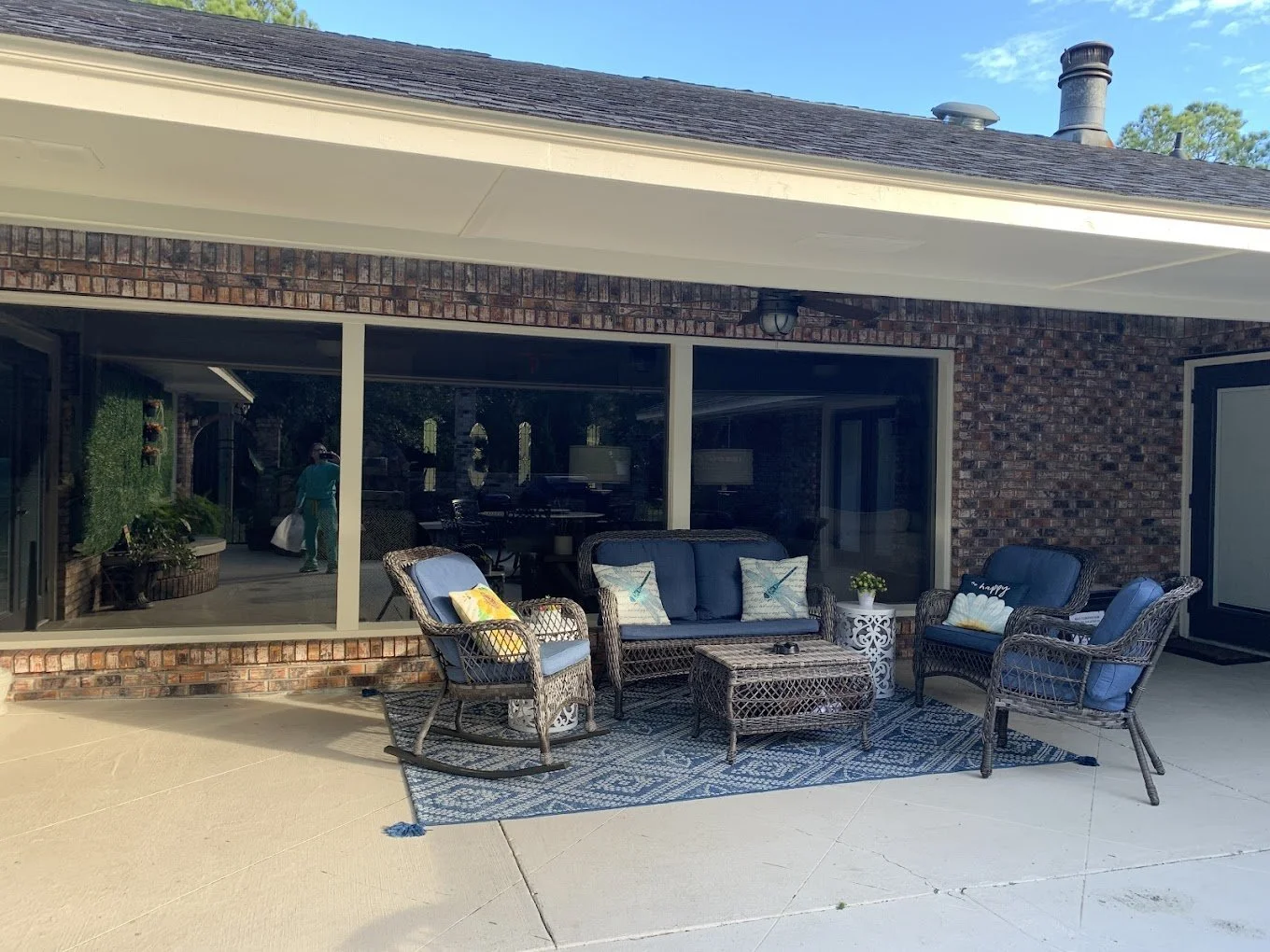 Outdoor patio area with wicker furniture including a sofa, chairs, and a coffee table, a patterned outdoor rug, against a brick house with large glass sliding doors and a reflection of a person inside.