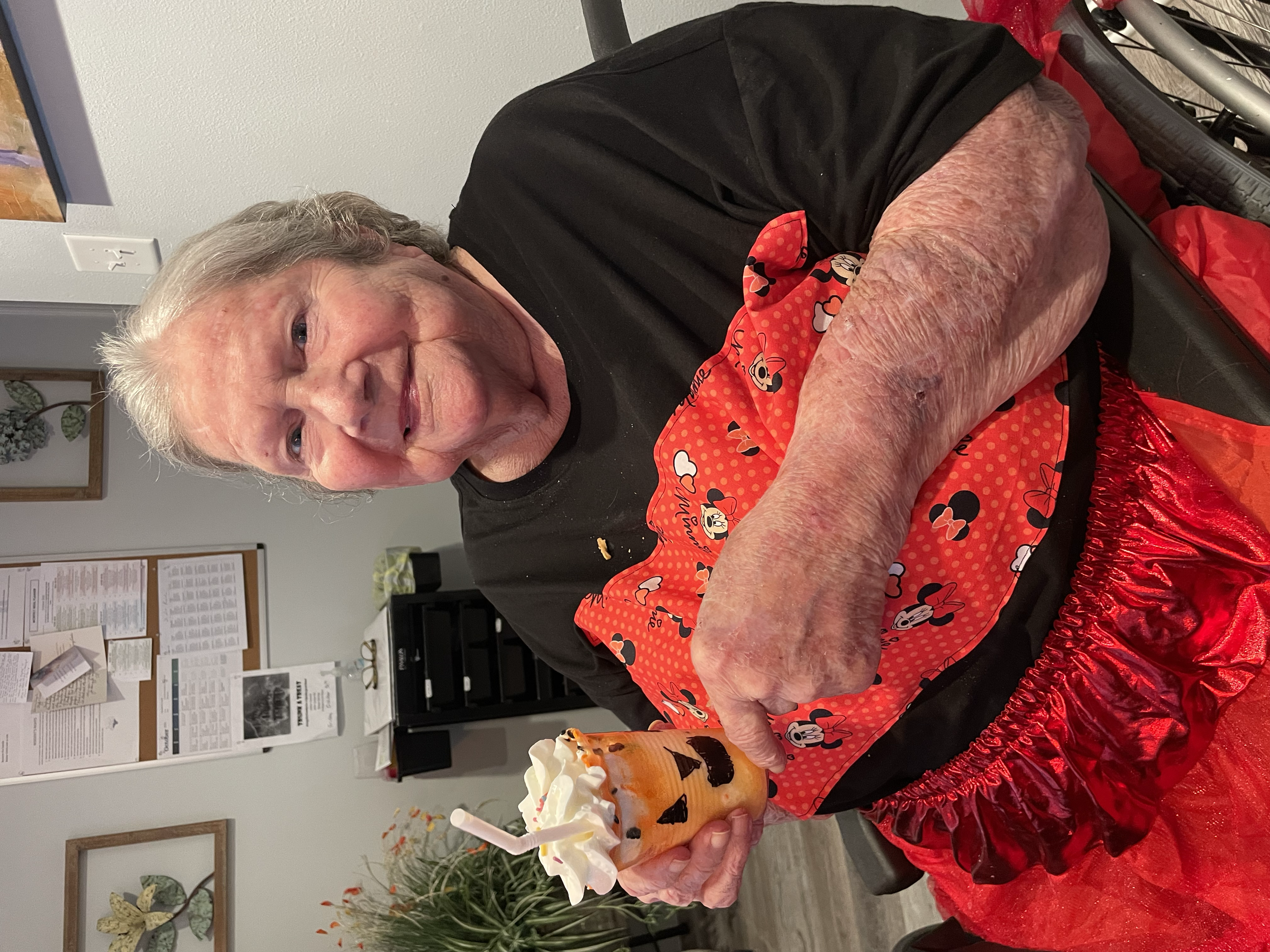 An elderly woman with gray hair smiling and holding a Halloween-themed drink decorated with pumpkin faces and whipped cream. She is wearing a Minnie Mouse themed scarf and is seated at a table with a red, shiny tablecloth. There are potted plants and