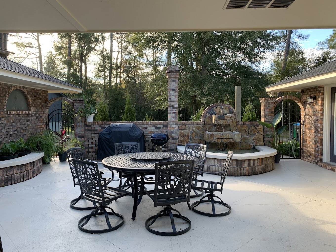 An outdoor patio area with a round dining table and six chairs, a covered grill, a water fountain made of stone, and lush greenery in the background, enclosed by brick walls and gates.
