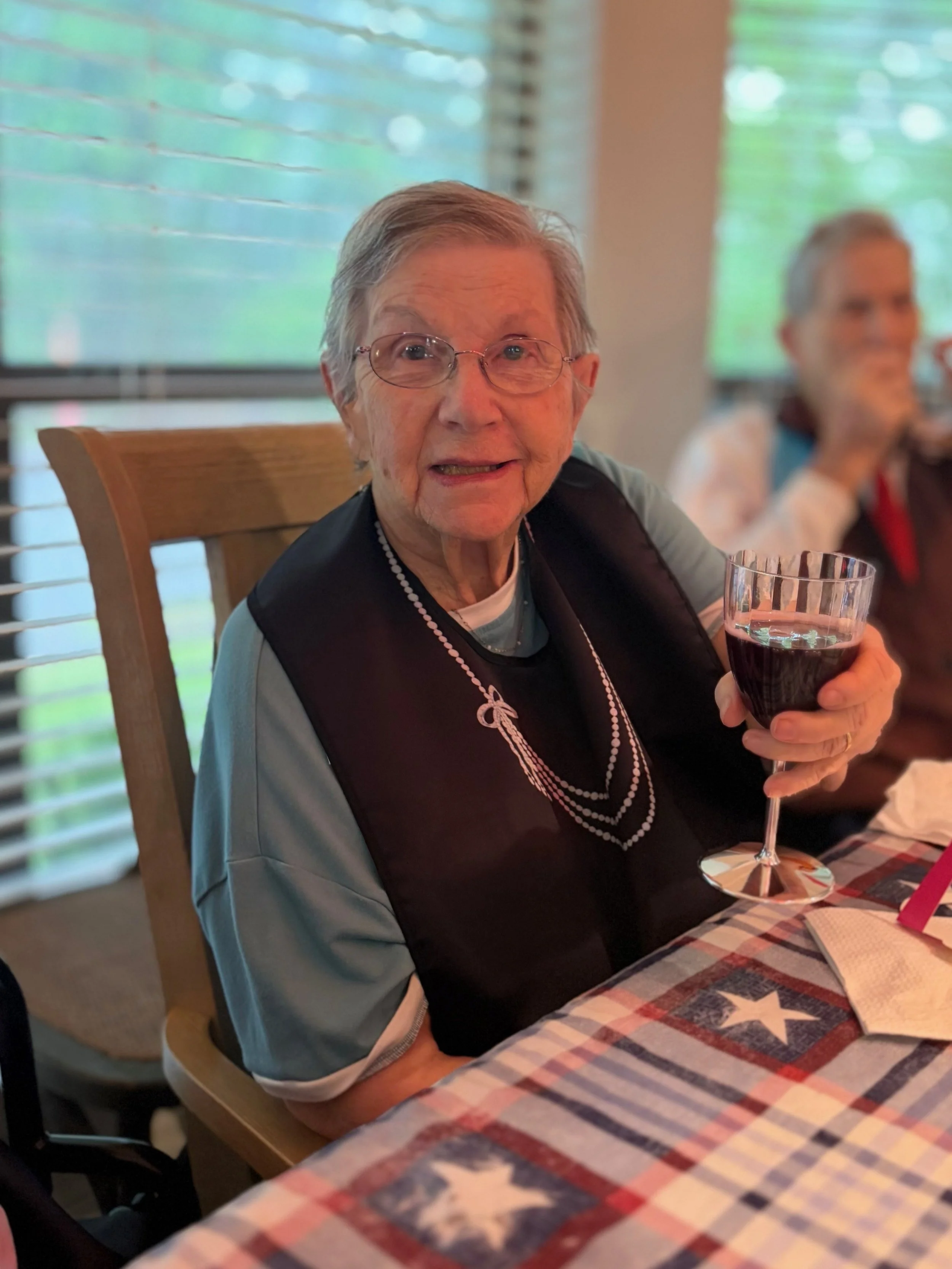 An elderly woman with glasses holding a glass of red wine, sitting at a table with an American flag patterned tablecloth in a dining area with window blinds.