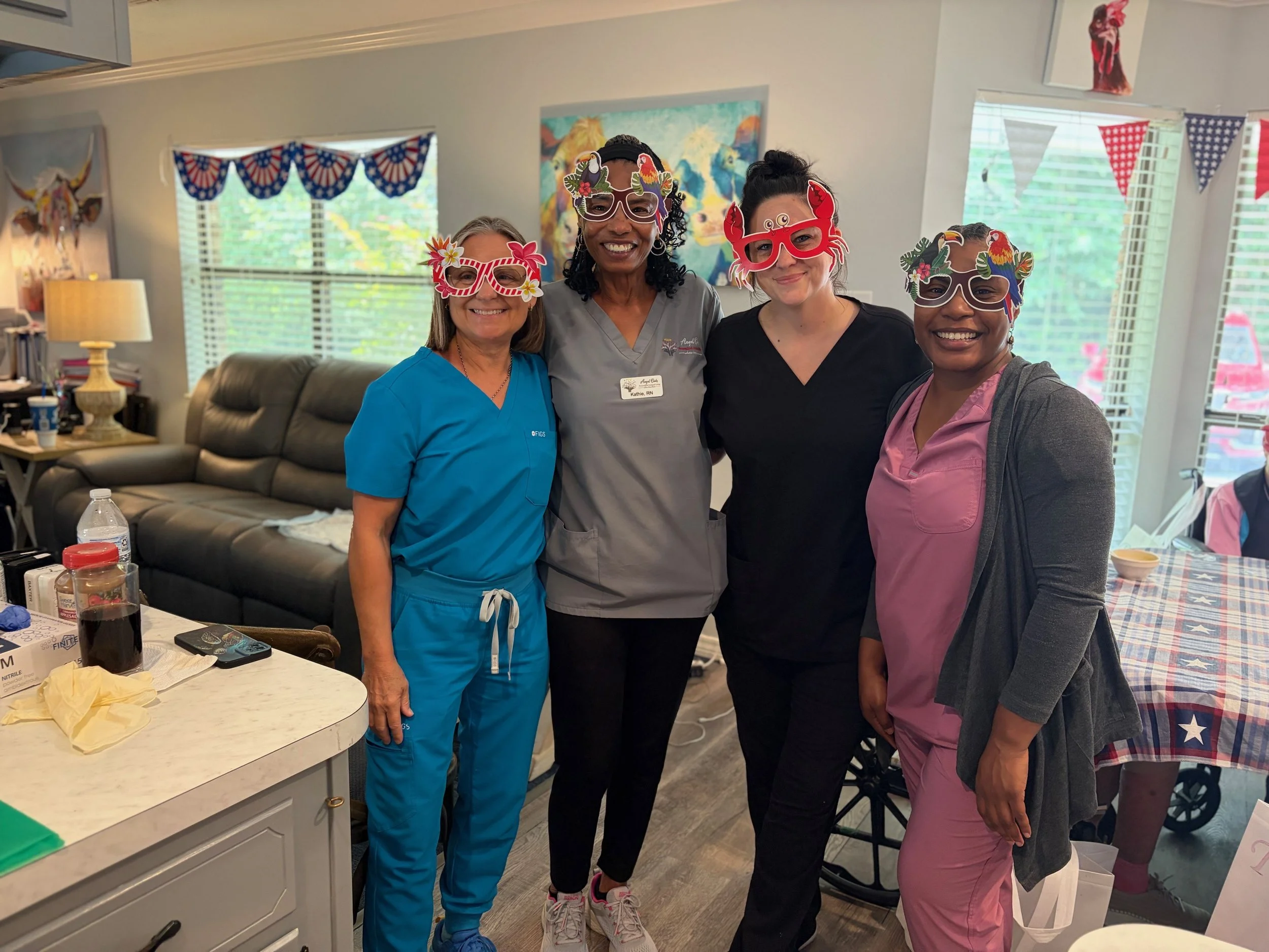 Four women in scrubs wearing colorful paper glasses with tropical and patriotic designs, smiling and standing close together in a decorated room with patriotic banners and artwork.