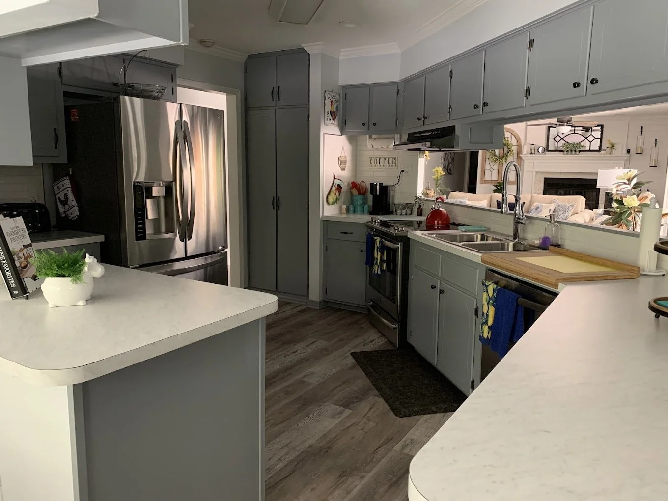Modern kitchen with gray cabinets, stainless steel refrigerator, marble countertops, and view into the living room with fireplace and beige sofa.