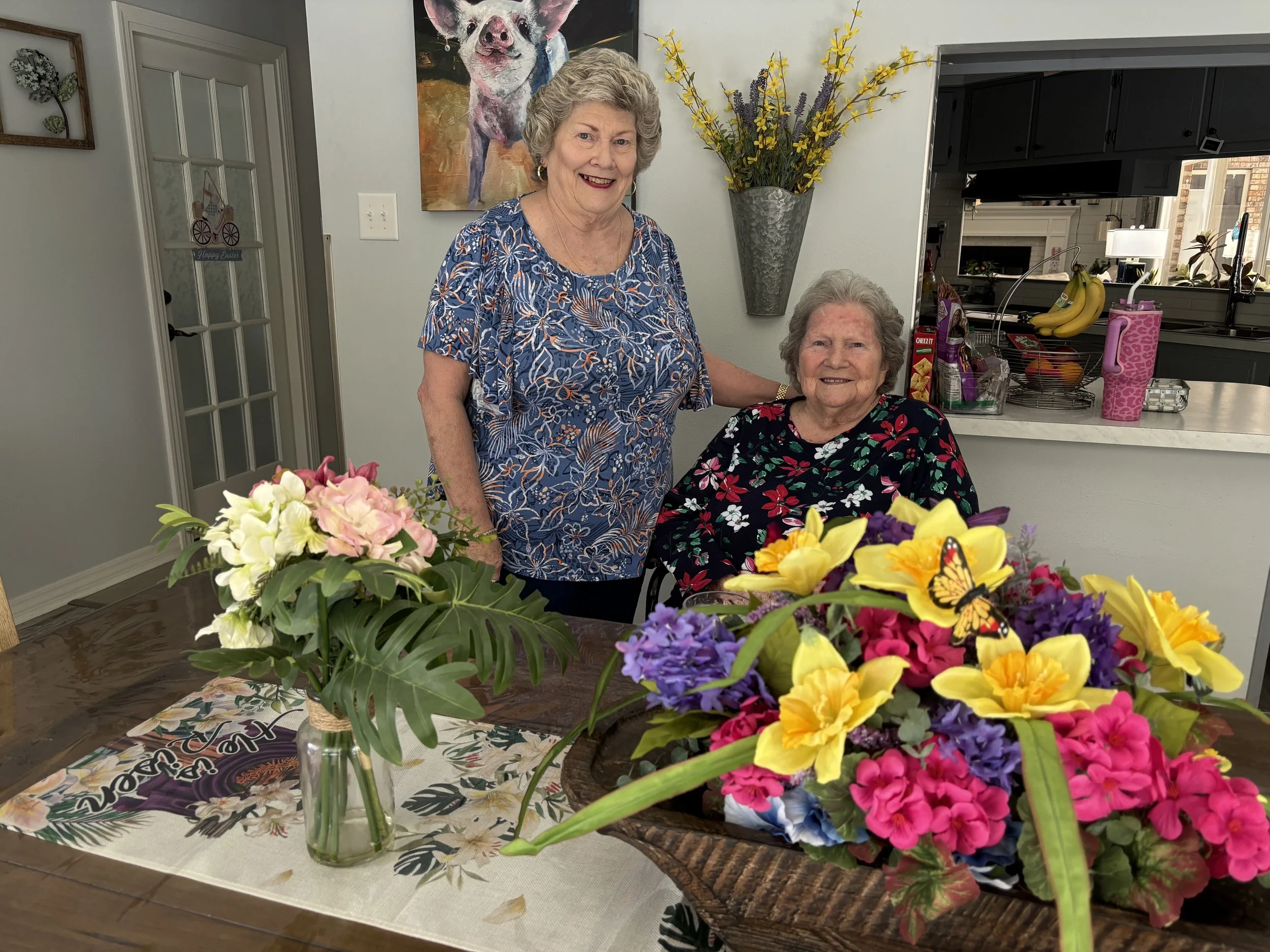 A woman stands next to another woman seated at a dining table with two large arrangements of colorful flowers in vases, in a bright kitchen. The standing woman is smiling and wears a blue patterned top; the seated woman also smiles and wears a floral