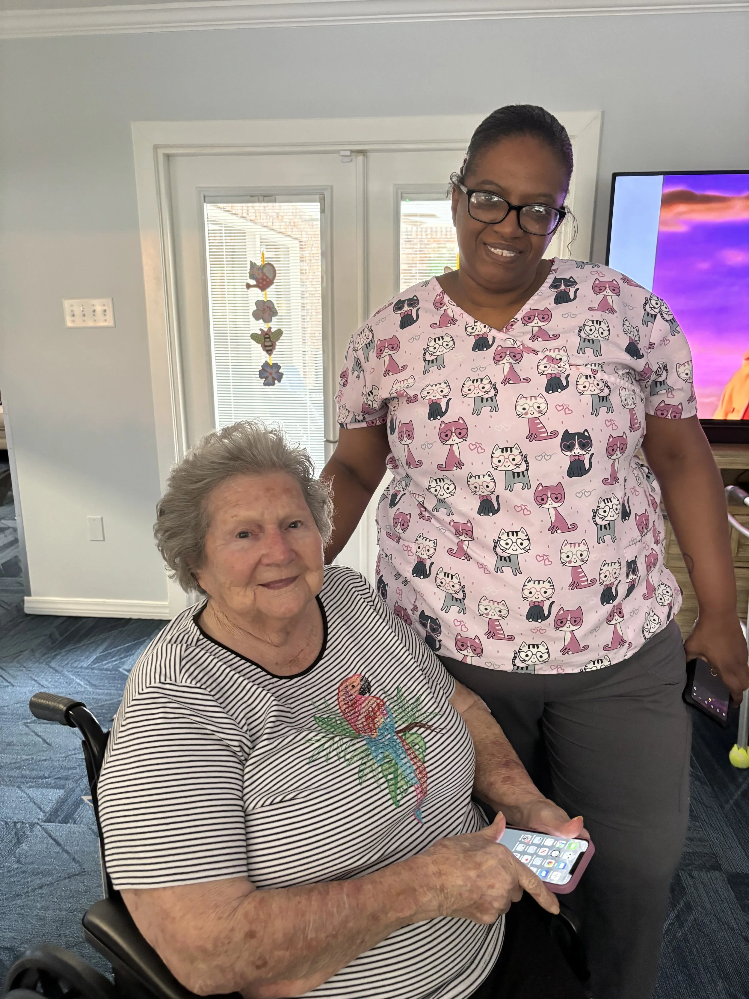 An elderly woman seated in a wheelchair holding a smartphone, with a younger woman standing beside her, both smiling inside a well-lit room with a TV and decorative hanging ornaments in the background.