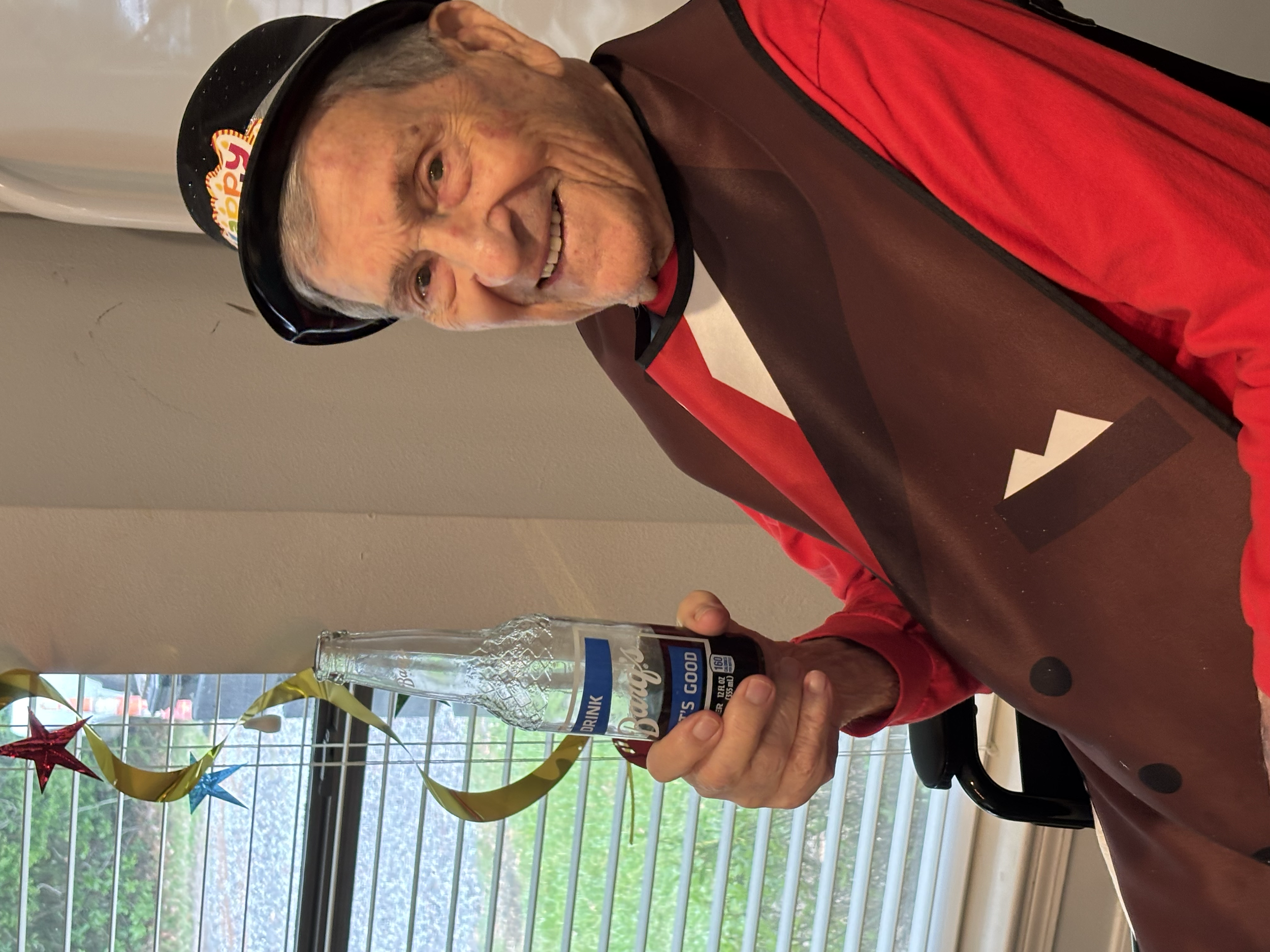 An elderly man smiling at the camera, holding a clear plastic cup of Bud Light beer with a straw. He is sitting indoors at a table decorated with colorful star-shaped balloons and streamers, in front of a window with blinds and trees outside.