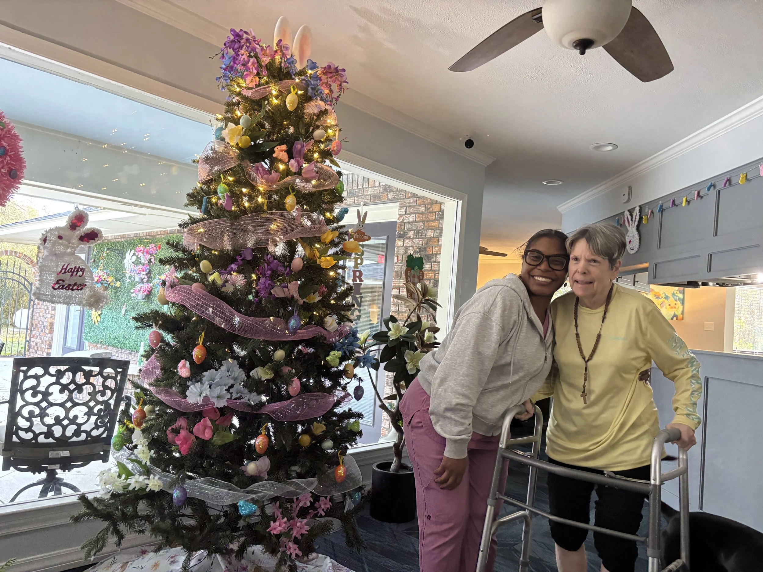A decorated Christmas tree with pink ribbons, bunny ears, and colorful ornaments next to two smiling women, one using a walker, inside a room with a ceiling fan and holiday decorations.