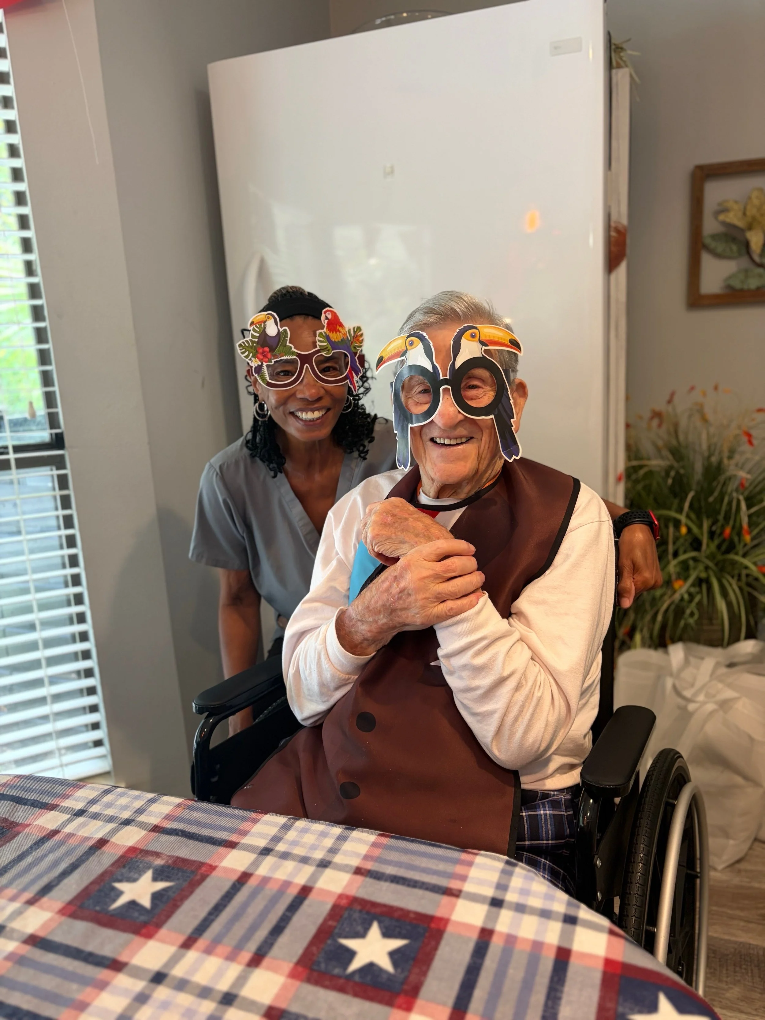 Two women wearing toucan-themed glasses celebrating together, with the elderly woman in a wheelchair and the younger woman standing behind her, both smiling.