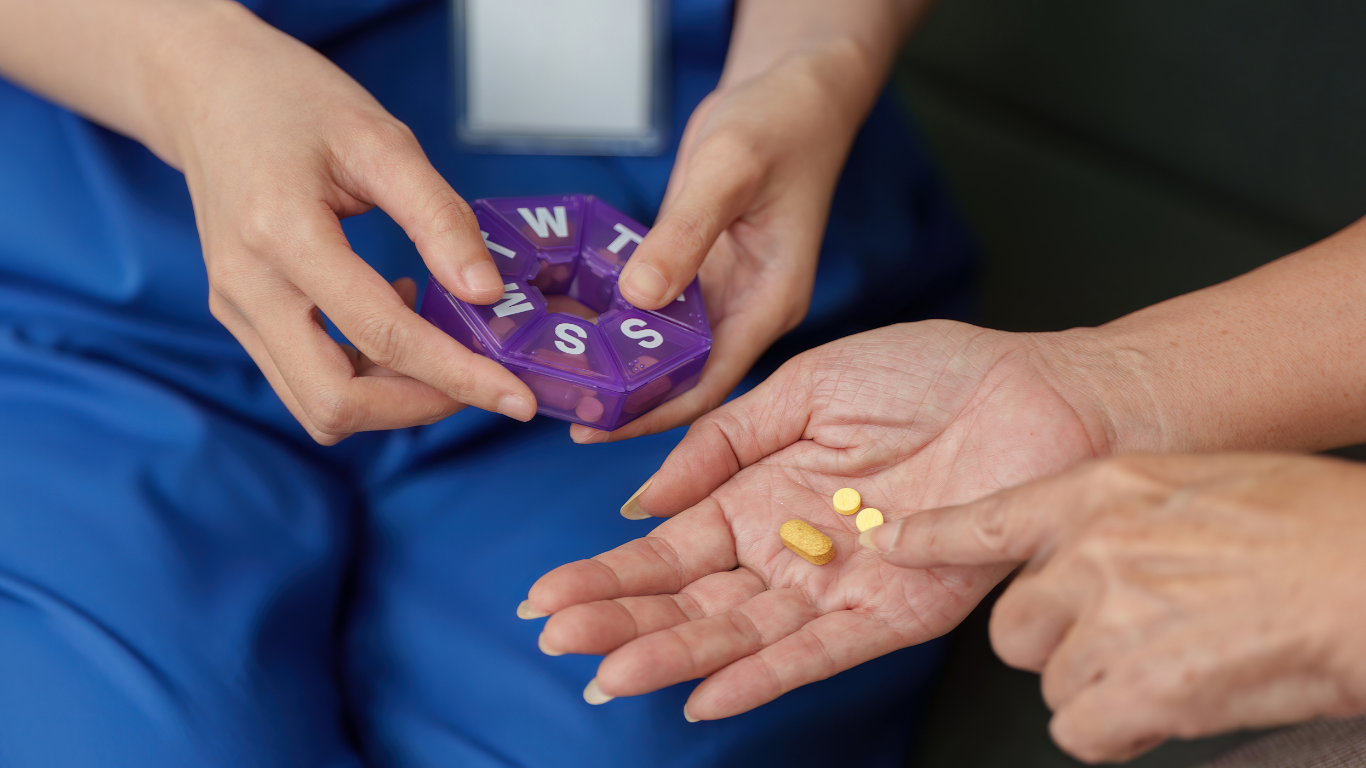 A healthcare professional handing a few yellow pills to a patient with an open palm, while the healthcare worker holds a purple pill organizer nearby.