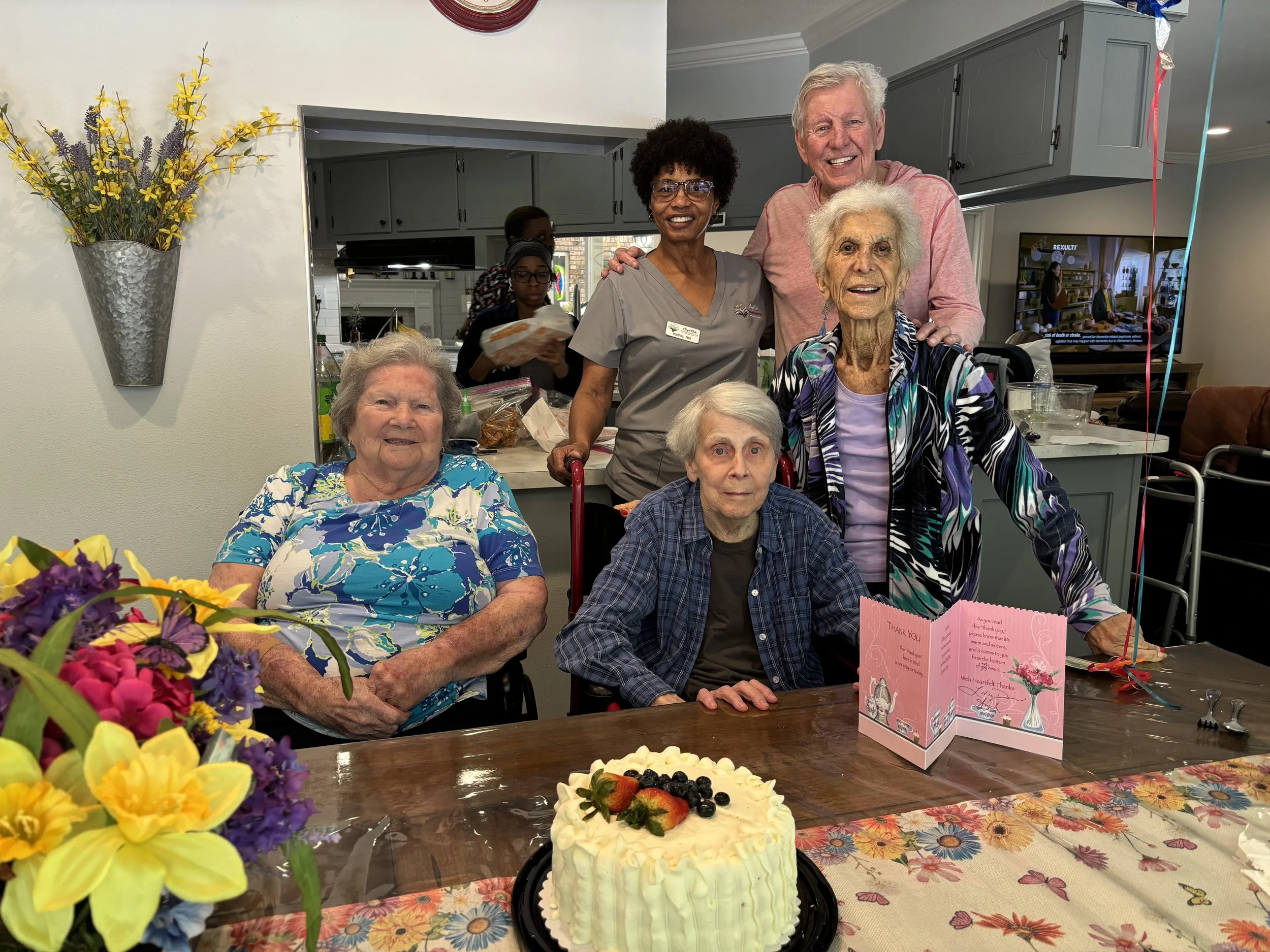 A group of elderly women and a man gathered around a decorated birthday cake with strawberries, blueberries, and strawberries on top. They are smiling and celebrating, with a flower arrangement and cards on the table. The background shows a kitchen a