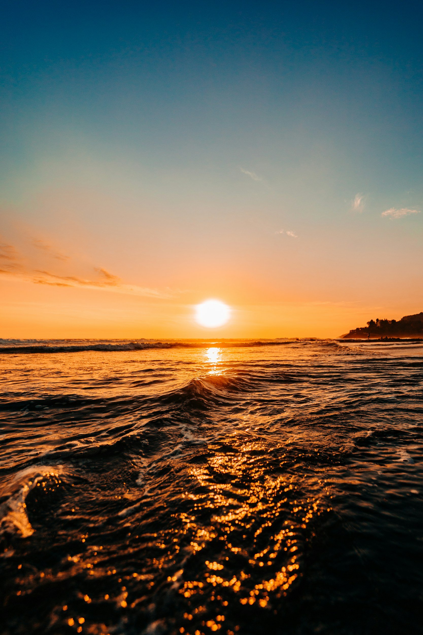 Sunset over the ocean with waves and a distant shoreline.