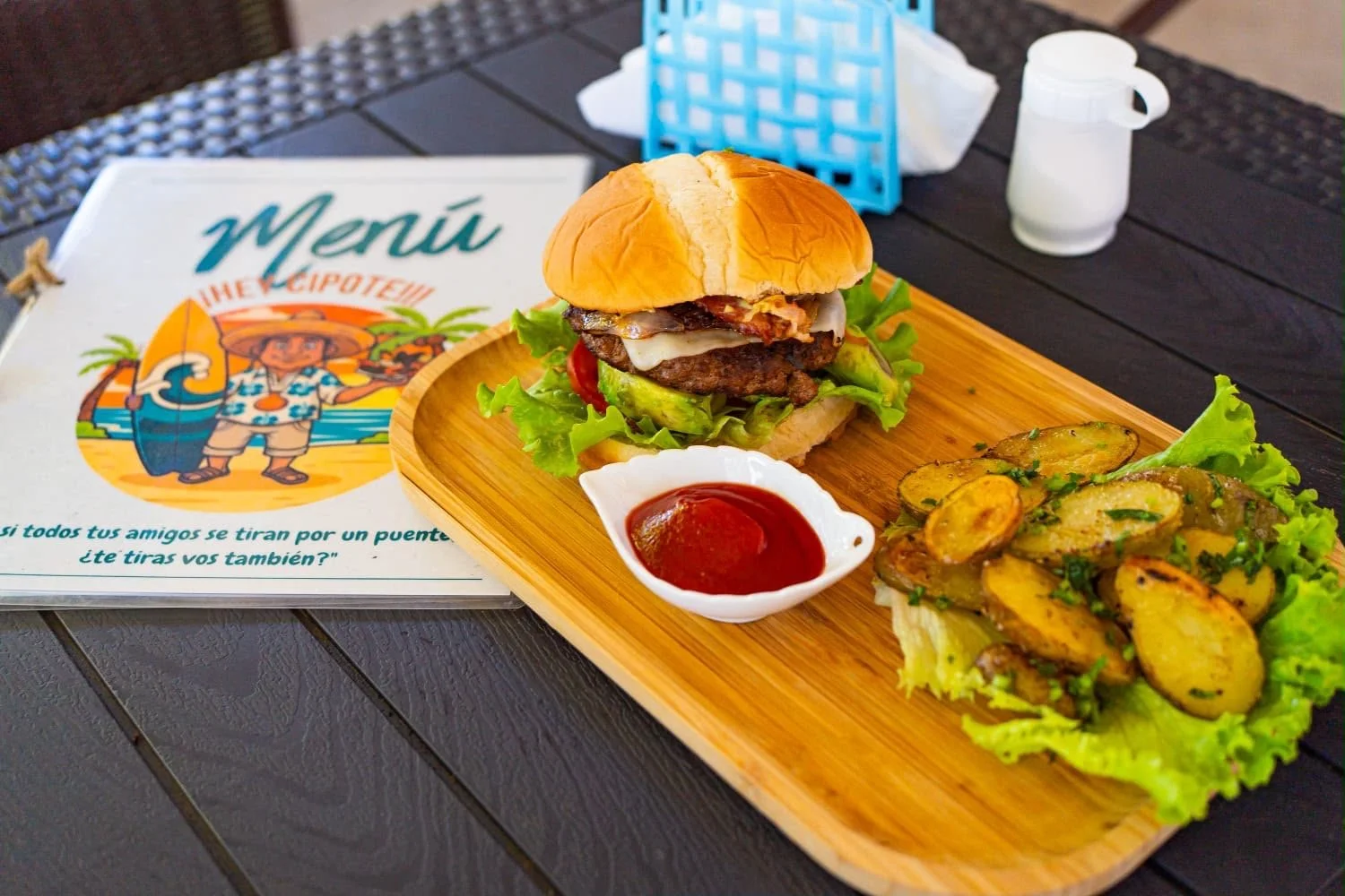 A hamburger with lettuce, tomato, cheese, and a beef patty on a bun, served with a side of roasted potatoes on a wooden tray with a small dish of ketchup and a side of lettuce. In the background, there is a restaurant menu with a cartoon figure of a man in a Hawaiian shirt holding a fishing rod and wearing a straw hat.