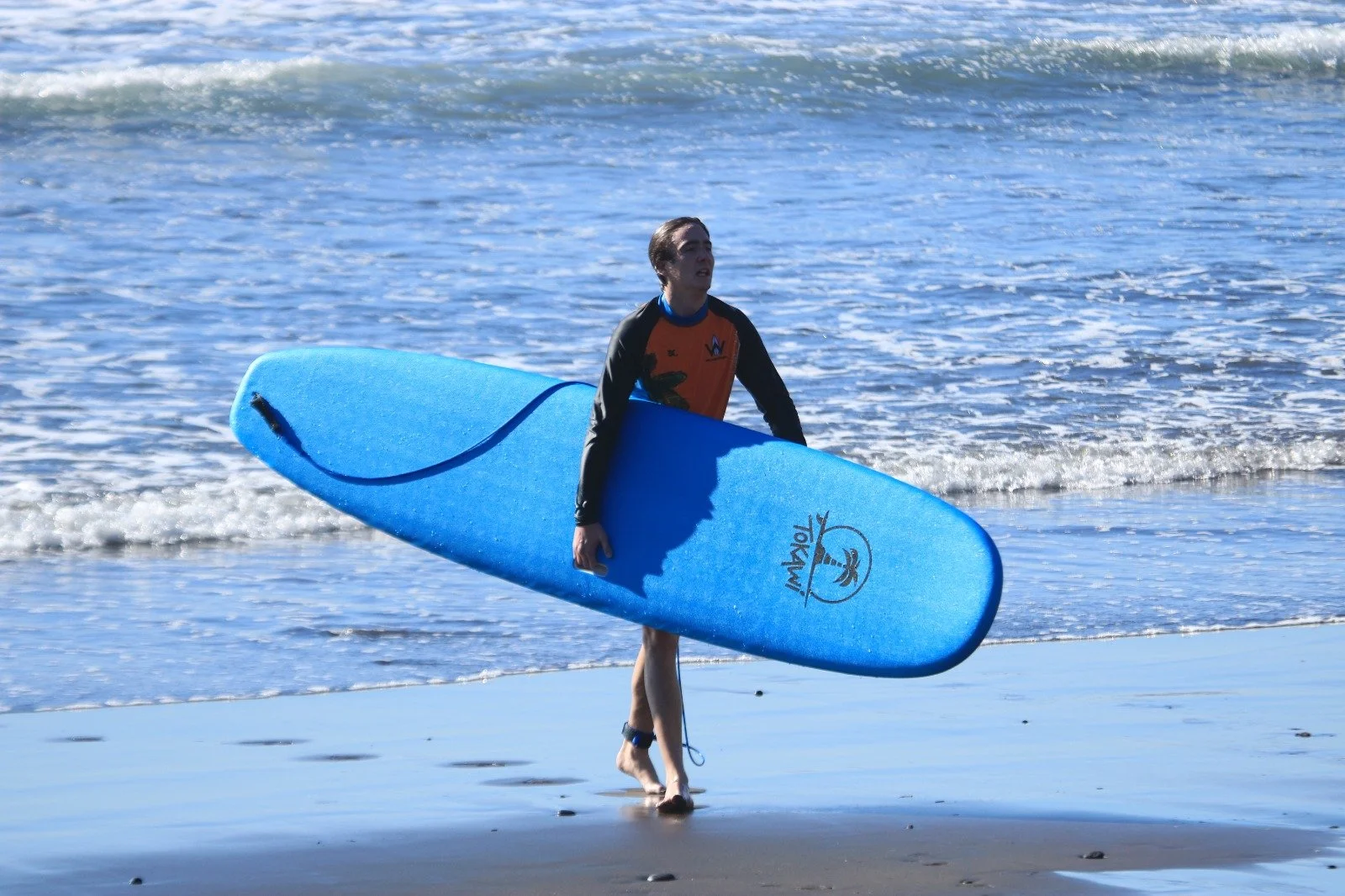 A young man carrying a blue surfboard while walking out of the ocean onto the sandy beach.