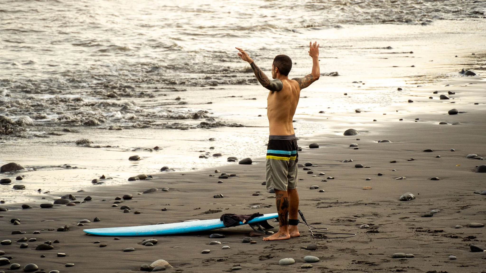 A man standing on a sandy beach with rocks, facing the ocean at sunset, next to a blue paddleboard.