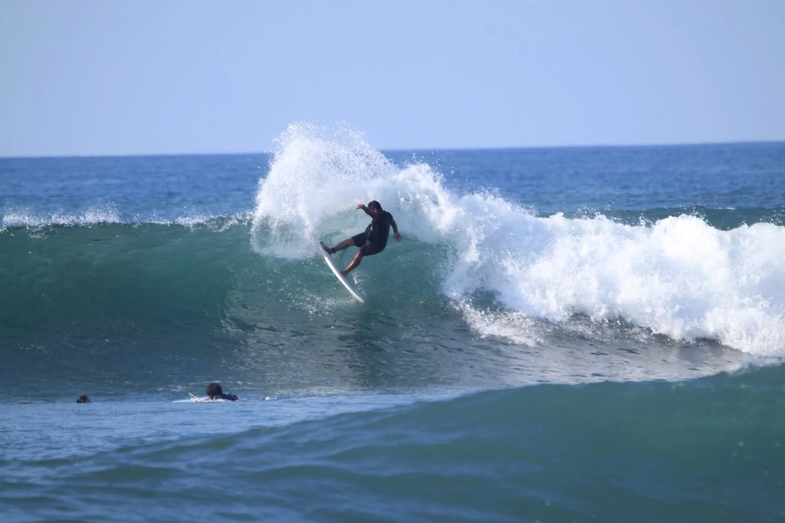 A person surfing on a wave with a splash of water behind them in the ocean, with a clear blue sky