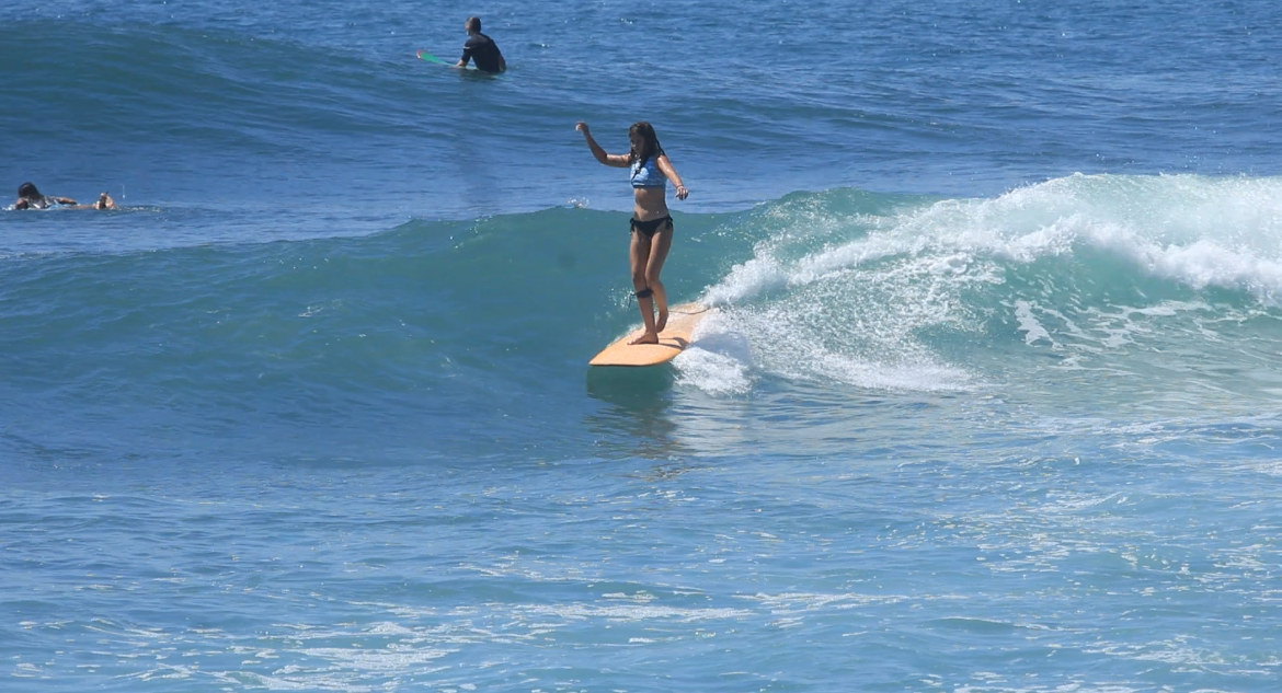 Woman surfing on a wave in the ocean, wearing a bikini, with other surfers in the background.