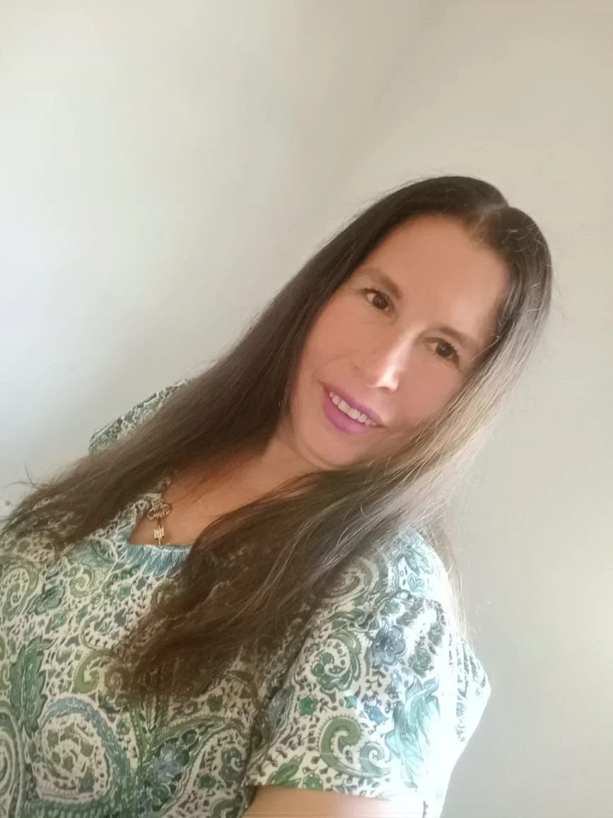 Mujer con cabello largo y oscuro, sonriendo, usando blusa con diseño de colores en tonos verdes y azules, con fondo de pared blanca.