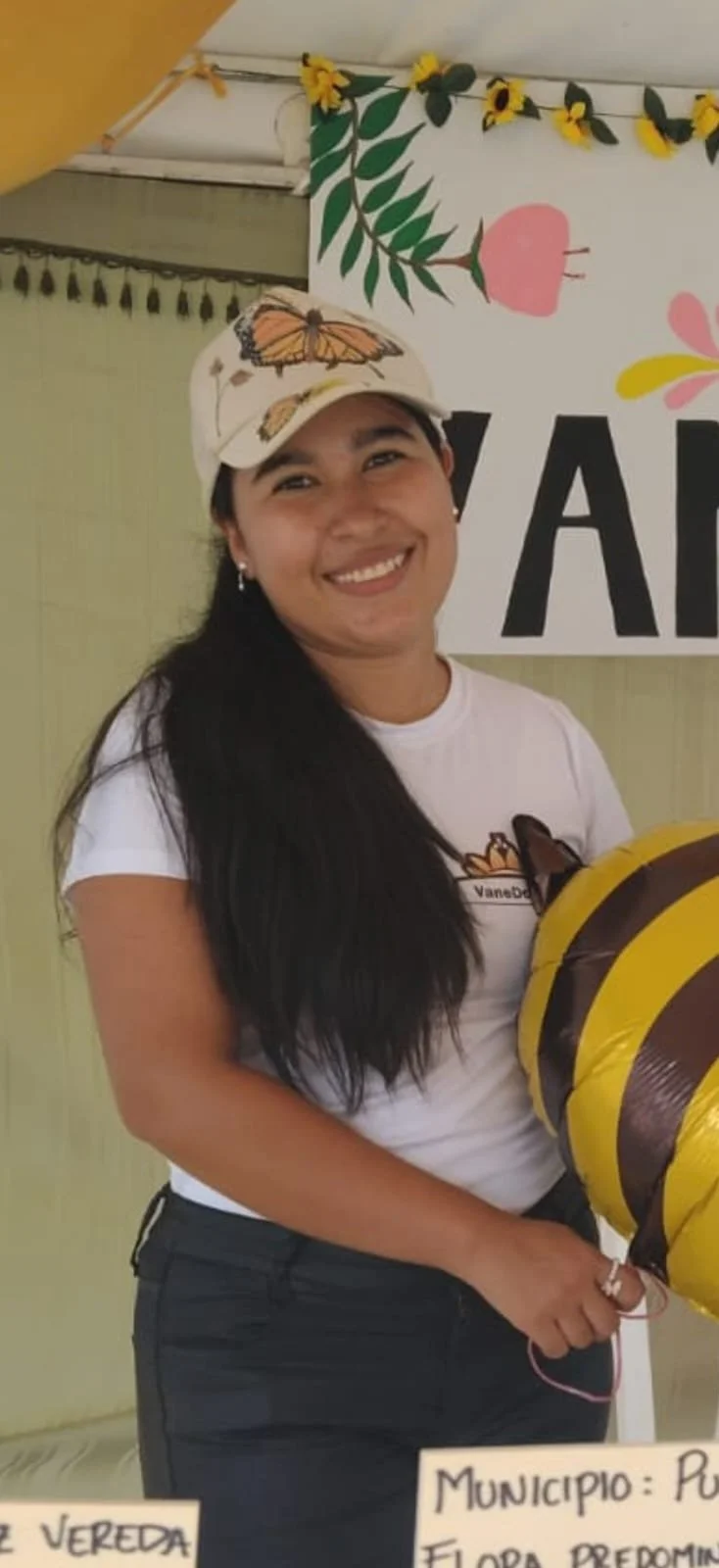 Mujer sonriendo, lleva gorra con mariposa, camiseta blanca y pantalón oscuro, en fondo decorado con dibujos de flores y hojas, en un evento municipal.