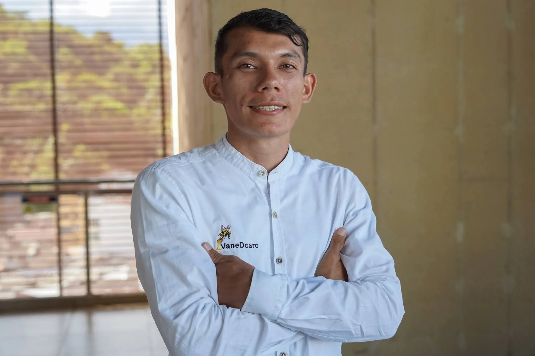 Retrato de un hombre joven de cabello oscuro, sonriendo y cruzado de brazos, vestido con una camisa blanca con bordado de una abeja y el texto 'VaneDcaro', en un interior con ventanas con persianas de madera y muro color beige.
