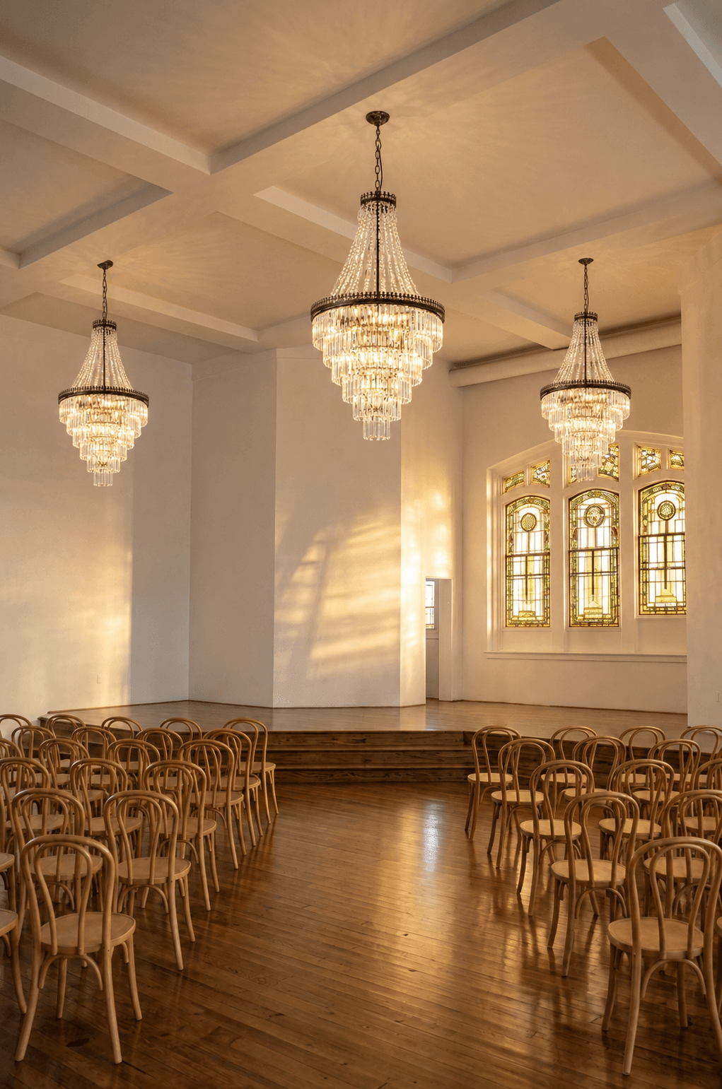 Elegant ceremony space at The Bradshaw with three large chandeliers hanging from a coffered ceiling, stained glass windows with intricate designs, and rows of wooden chairs on a polished hardwood floor, ready for an event.