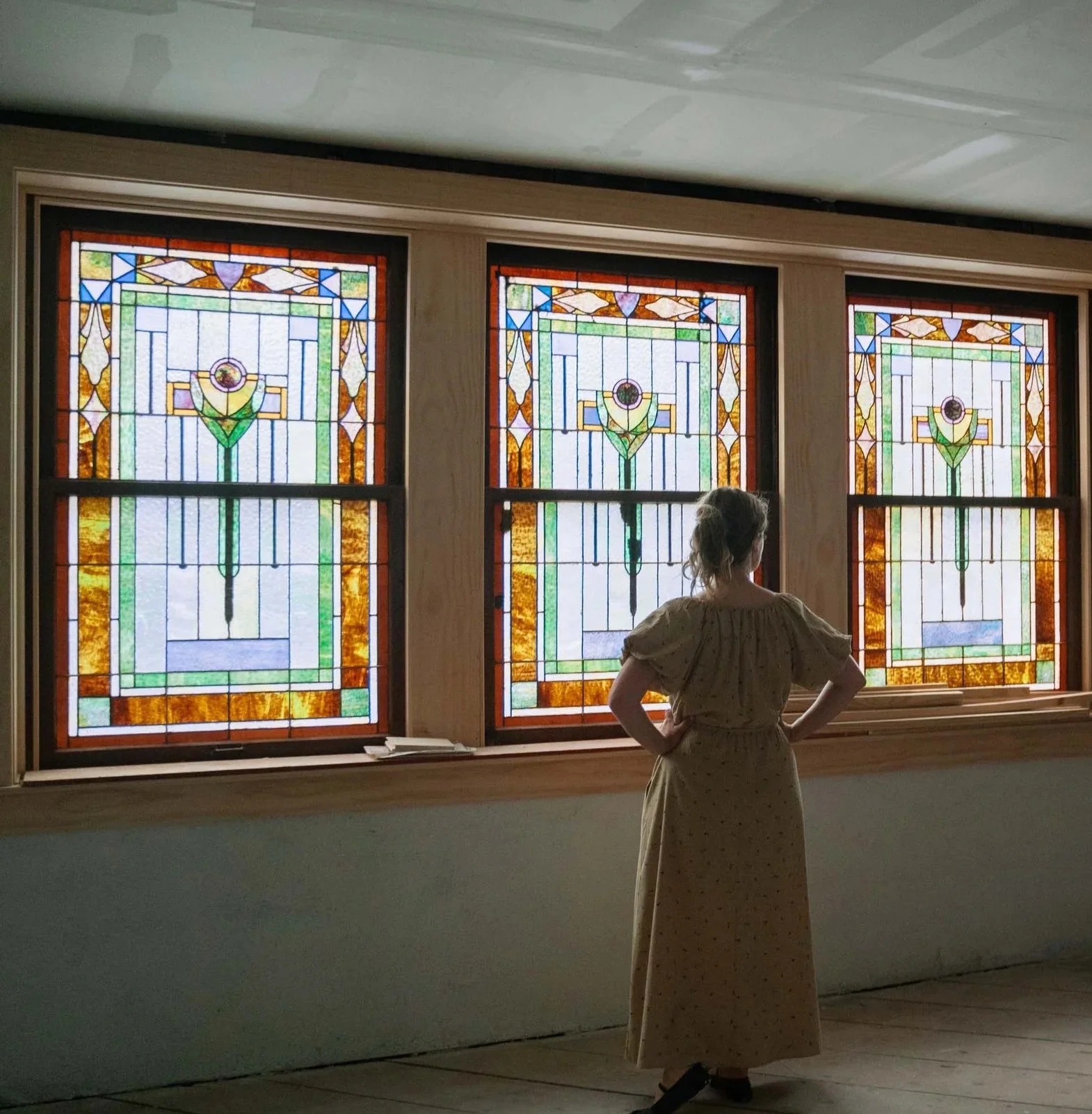 A woman in a dress stands looking at three stained glass windows with colorful geometric designs in The Bradshaw historic wedding venue in Eudora, Kansas.