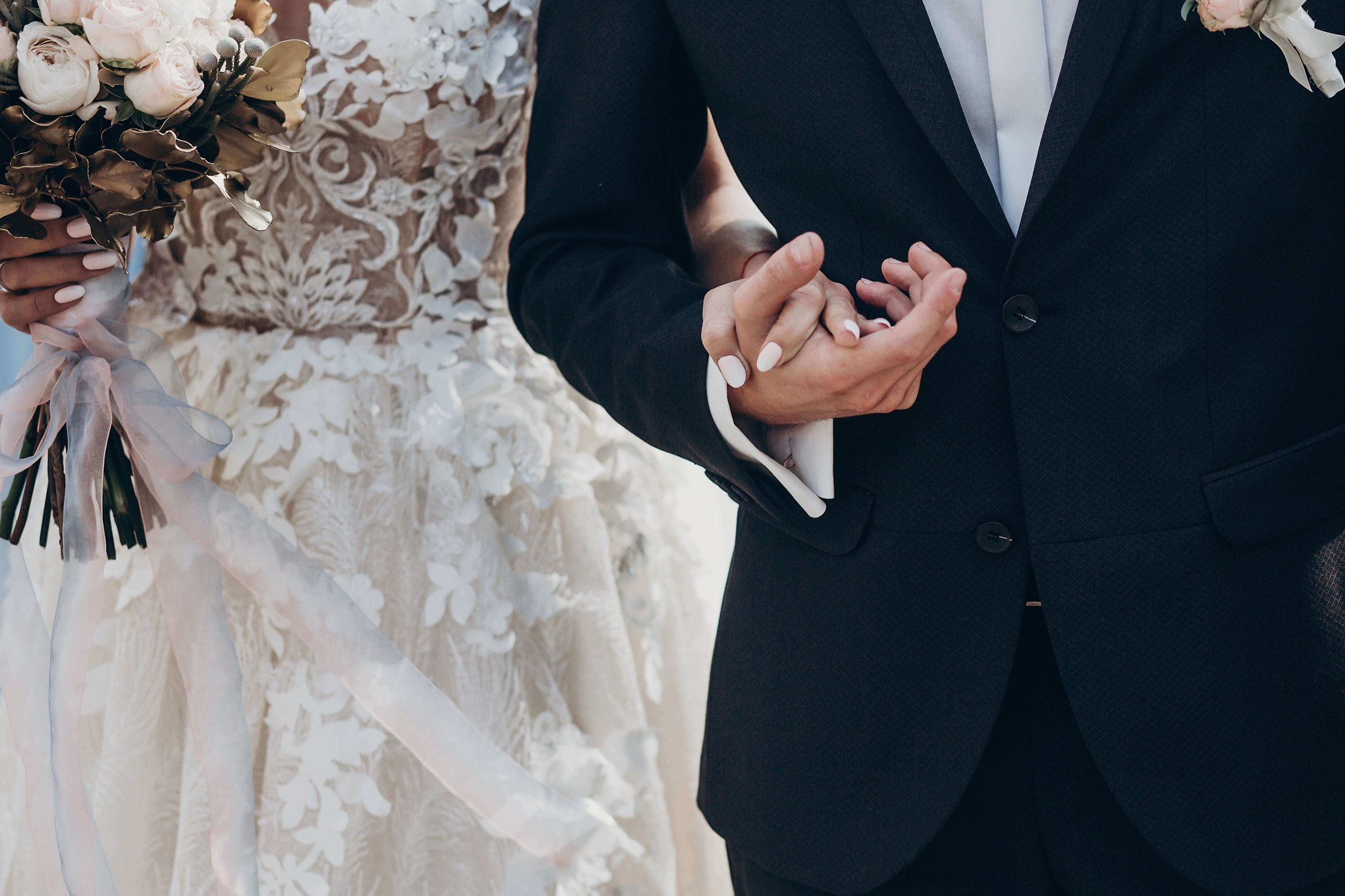 A couple holding hands, dressed in wedding attire, with the bride holding a bouquet flowers and the groom wearing a dark suit.