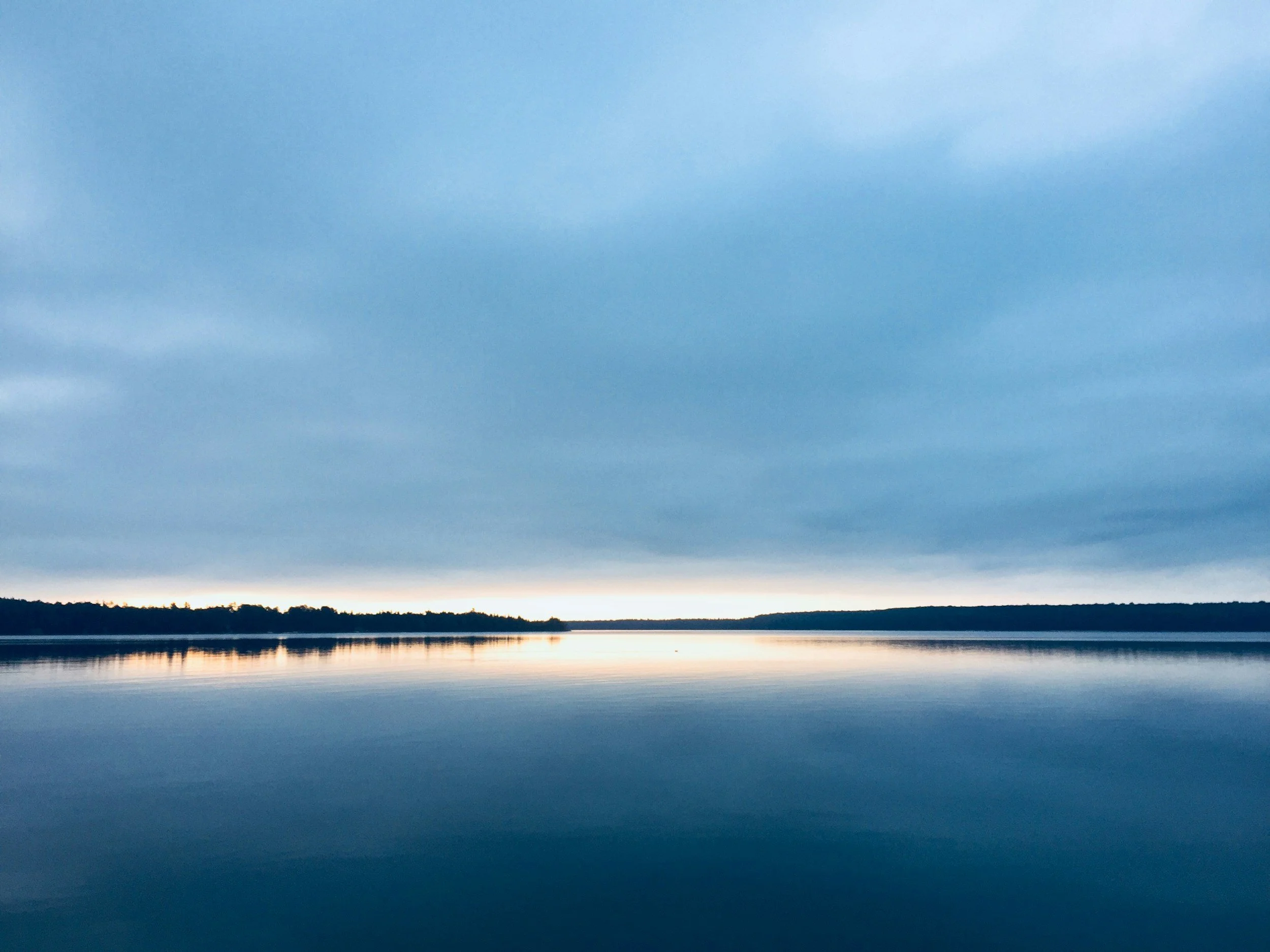 A wide view of a lake at dusk with calm water reflecting the cloudy sky and a silhouette of a forested shoreline in the distance.