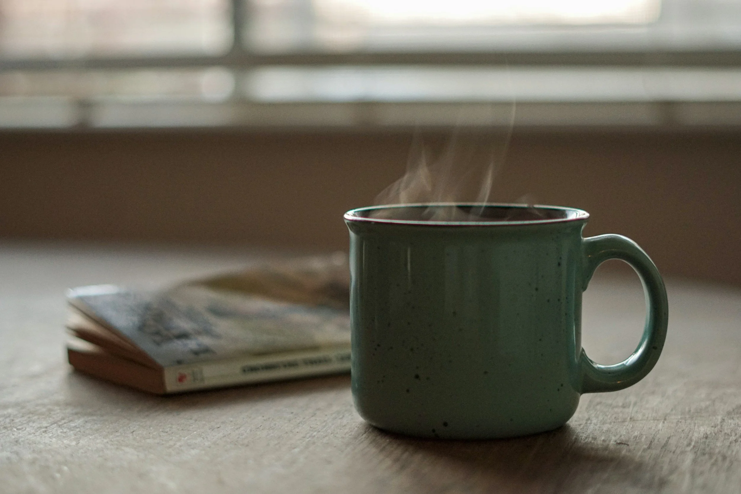 A steaming green ceramic mug sitting on a table with a book and a magazine nearby, with blurred window blinds in the background.