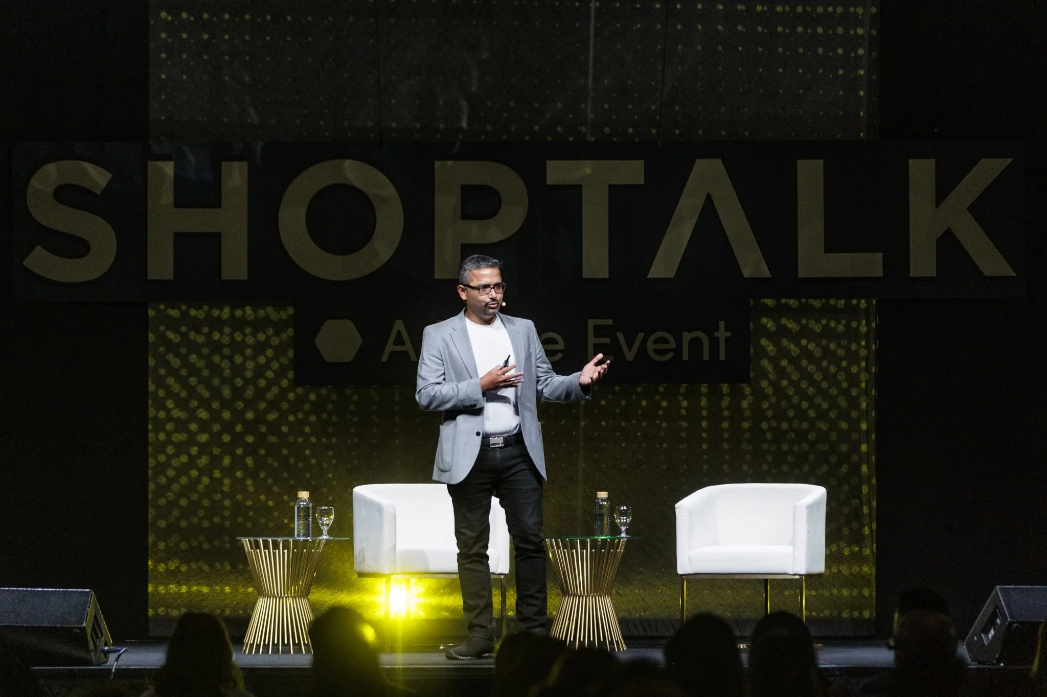 A man in a gray blazer and black pants is speaking on stage at a conference, with a large sign behind him reading 'SHOPTALK'. There are two white chairs and a small table with water bottles and glasses on the stage.