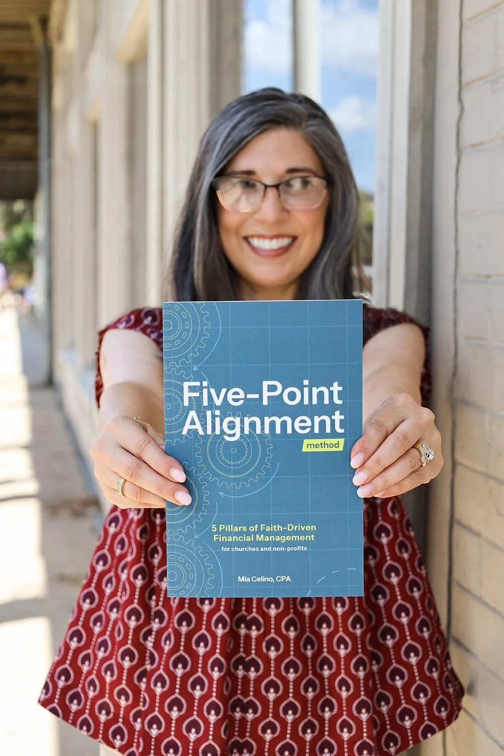 Mia Celino, CPA, holding the Five-Point Alignment Method booklet. She is standing outside next to a brick wall with a wooden building and a clear sky in the background.