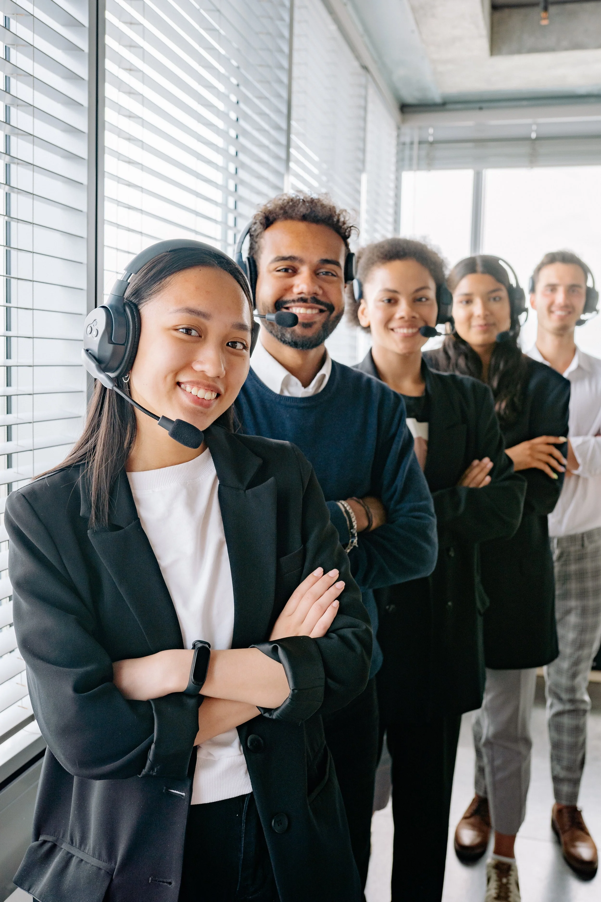 Five diverse customer service representatives standing in a row, wearing headsets with microphones, smiling, indoors with large windows and blinds.