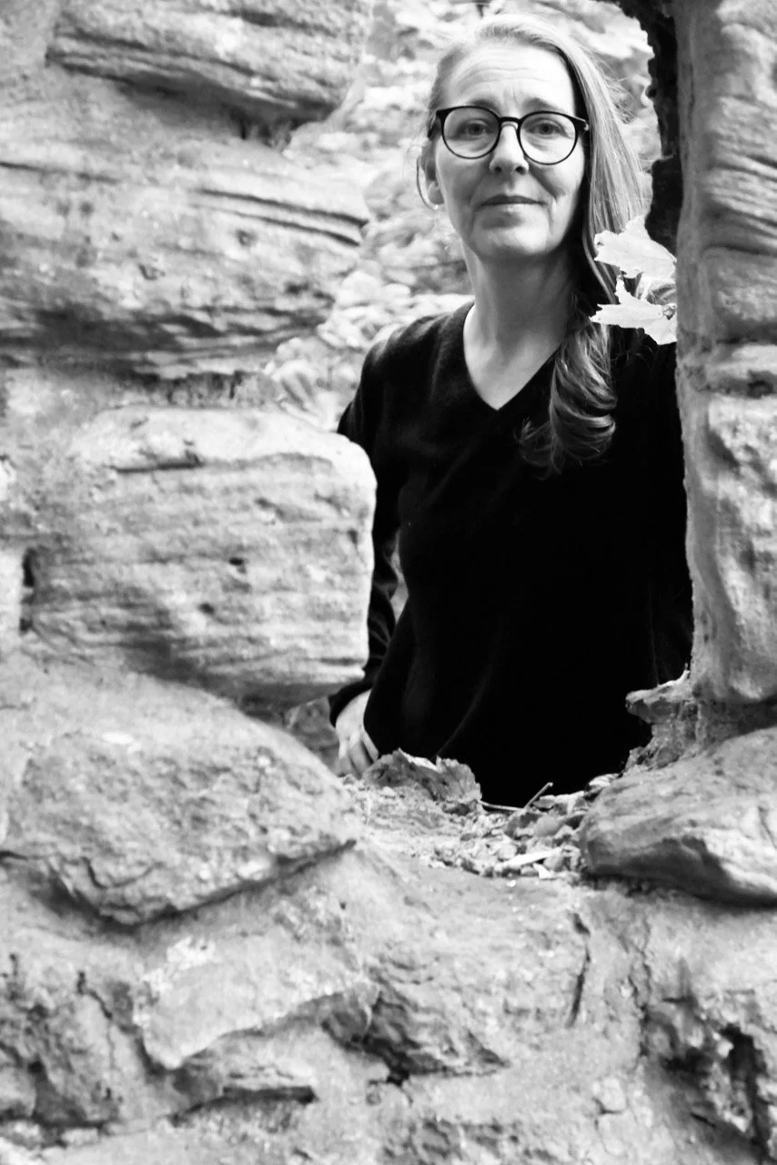 A woman with glasses and long hair, wearing a black shirt, standing among rocky formations, peering through a gap in the rocks.