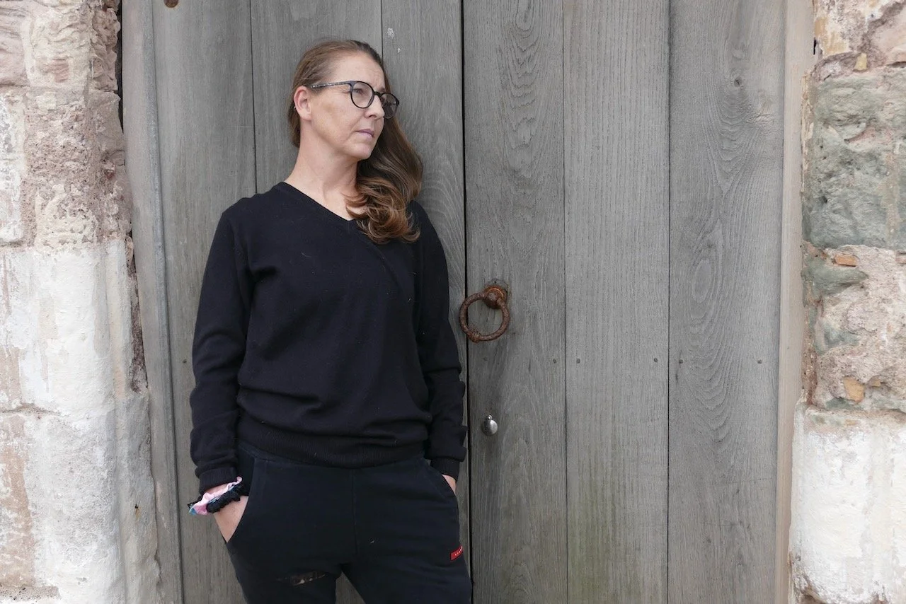 A woman with glasses and long brown hair standing in front of a gray wooden door with a rusty ring handle, wearing a black sweatshirt and black pants with hands in pockets.