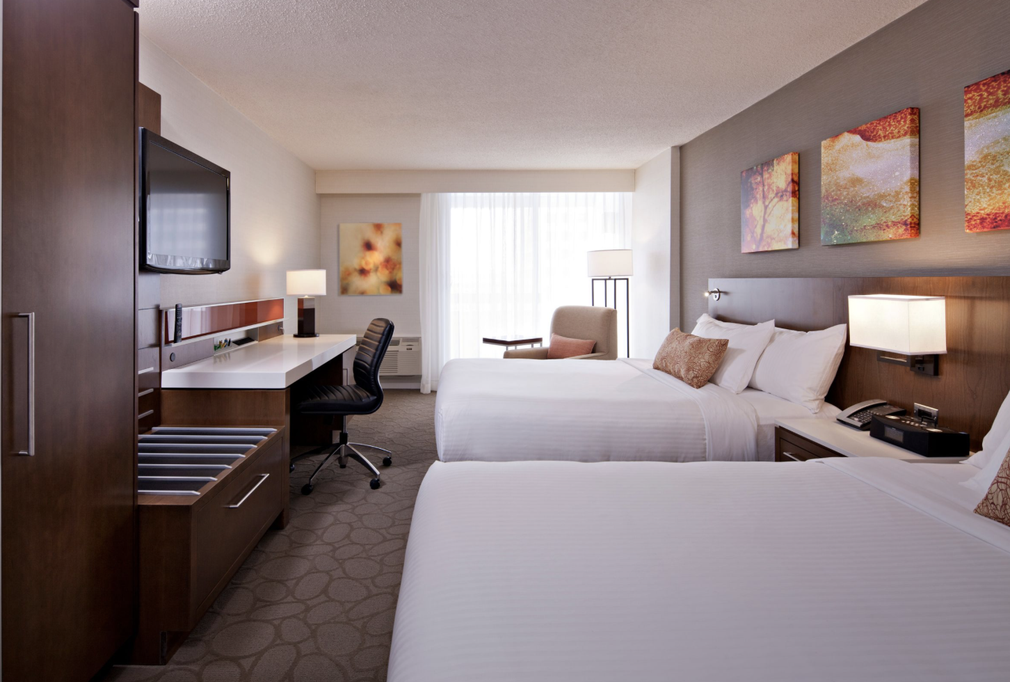 Hotel room with two beds, a desk, a chair, a flat-screen TV on the wall, and decorative wall art.
