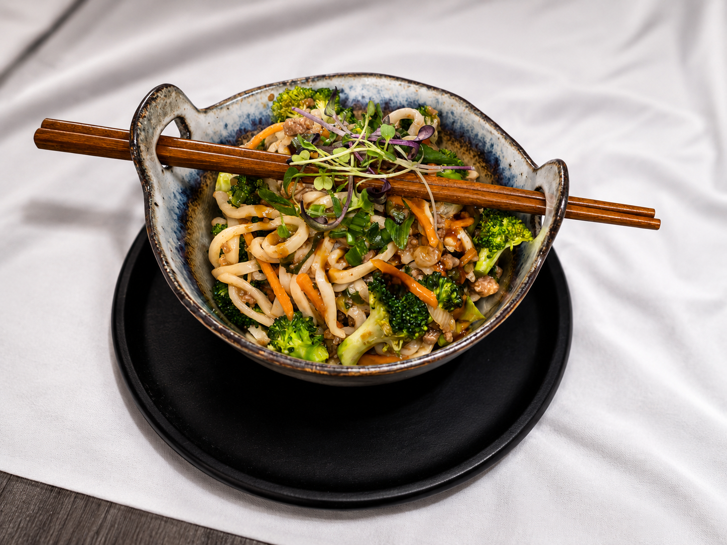 A bowl of Asian stir-fry with noodles, broccoli, carrots, and green vegetables, topped with microgreens, with wooden chopsticks resting on top, on a black round tray, on a white tablecloth.