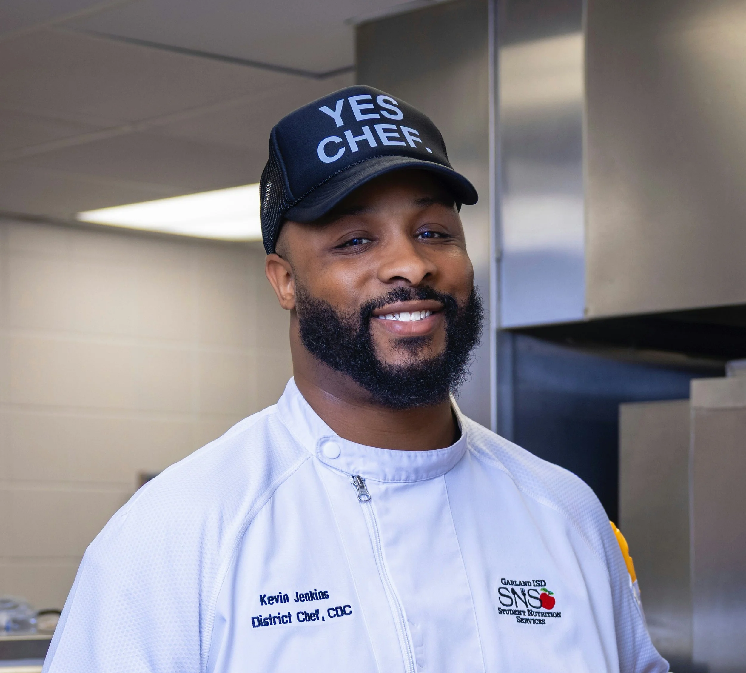A man wearing a black cap with white text that says "YES CHEF" and a white jacket with embroidered name and titles, smiling, with kitchen equipment and cabinets in the background.