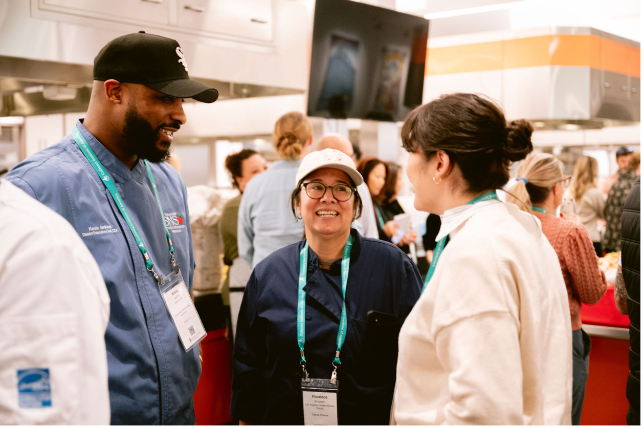 Three people engaged in a conversation at a conference or event, with a busy background showing other attendees and kitchen area.