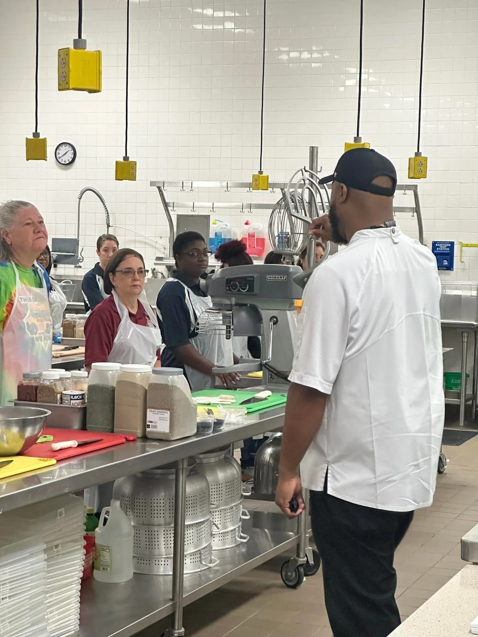 People in a professional kitchen or food preparation area, wearing aprons and hair coverings, with cooking supplies and equipment visible on the stainless steel counter.