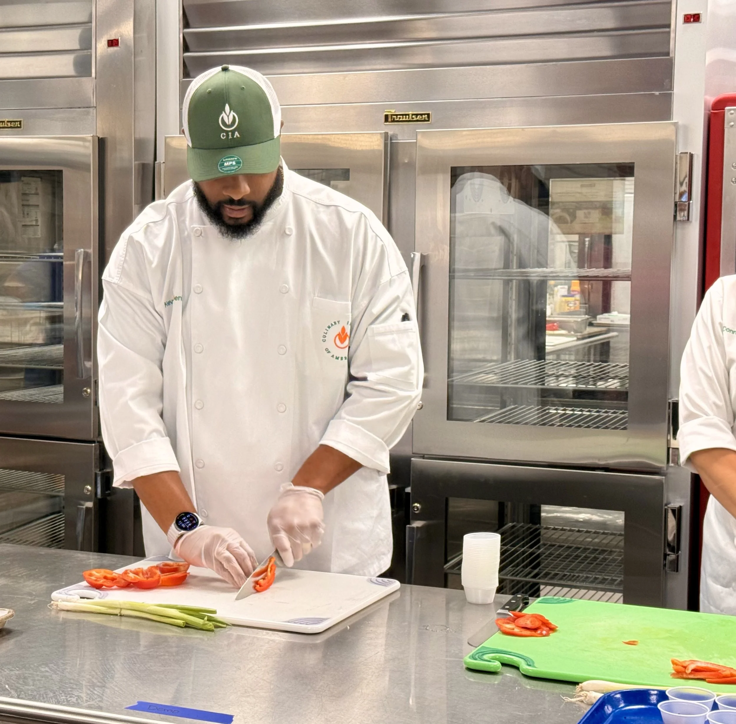 Chef chopping red bell peppers on a white cutting board in a commercial kitchen, with green onions and various containers on the gray countertop.