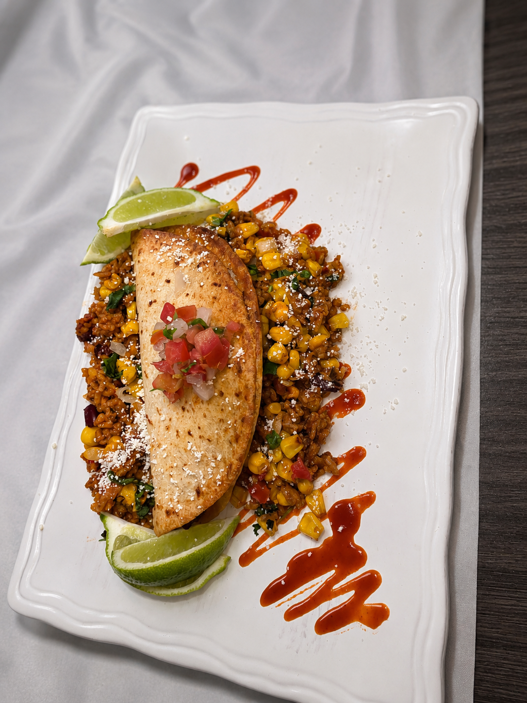 Seared fish fillet served on a bed of seasoned rice and corn salad, garnished with lime wedges, diced tomato and onion pico de gallo, drizzled with red and orange sauces, on a white decorative plate.