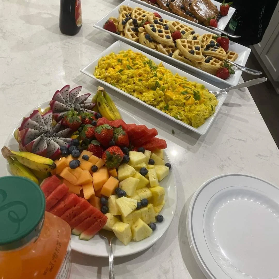 A table set with various breakfast foods, including a fruit platter with watermelon, cantaloupe, pineapple, strawberries, blueberries, dragonfruit, and bananas; a plate of scrambled eggs garnished with green herbs; a rectangular tray of waffles toppe