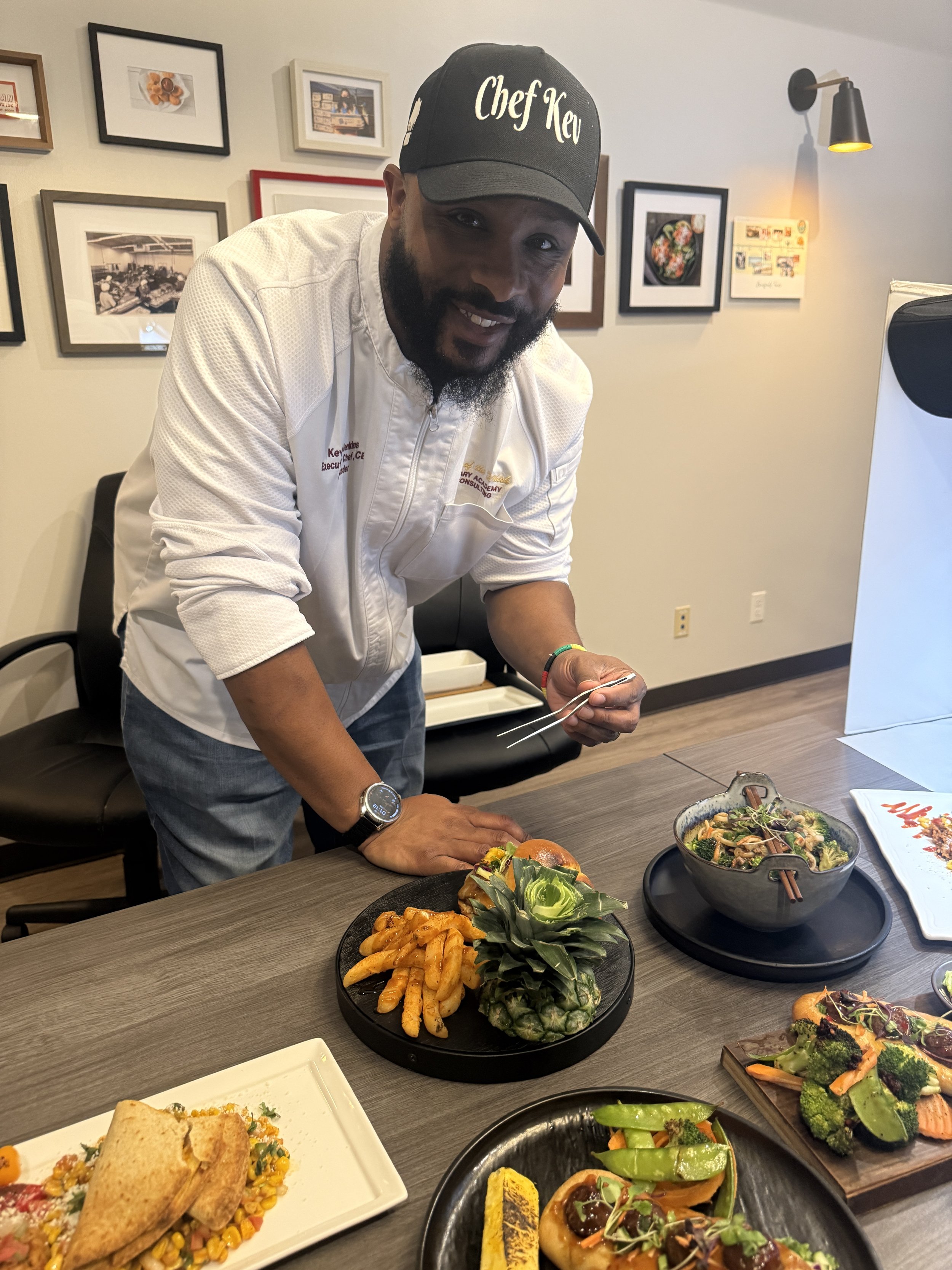 A man wearing a black cap labeled 'Chef Key' and a white chef jacket, smiling and holding a fork, stands at a table filled with various Asian dishes, in a room decorated with framed pictures on the wall.