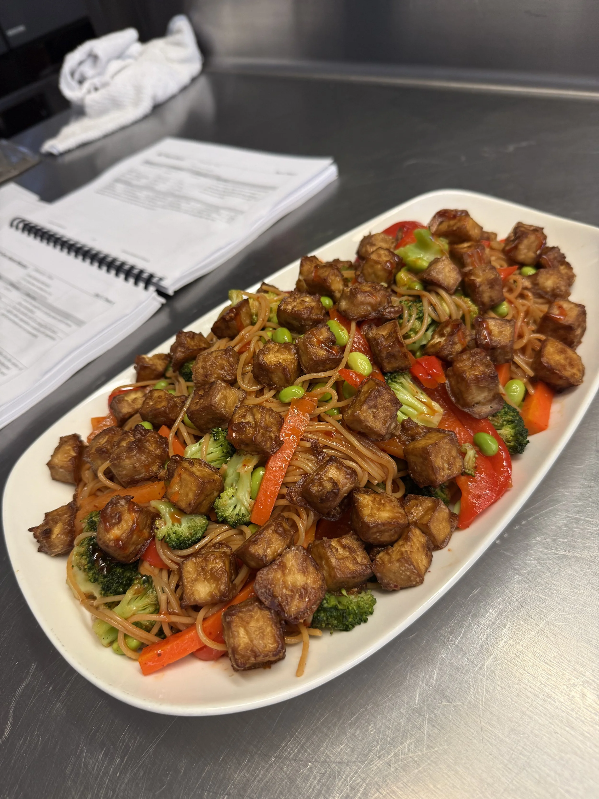 Plate of stir-fried noodles with tofu and vegetables on a stainless steel countertop in a kitchen.