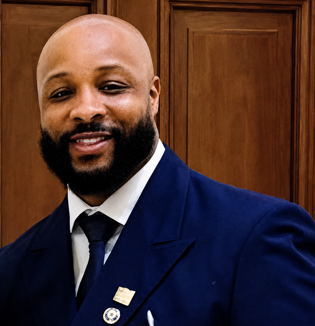 A man with a bald head and beard dressed in a blue suit and tie, standing in front of wooden cabinets.